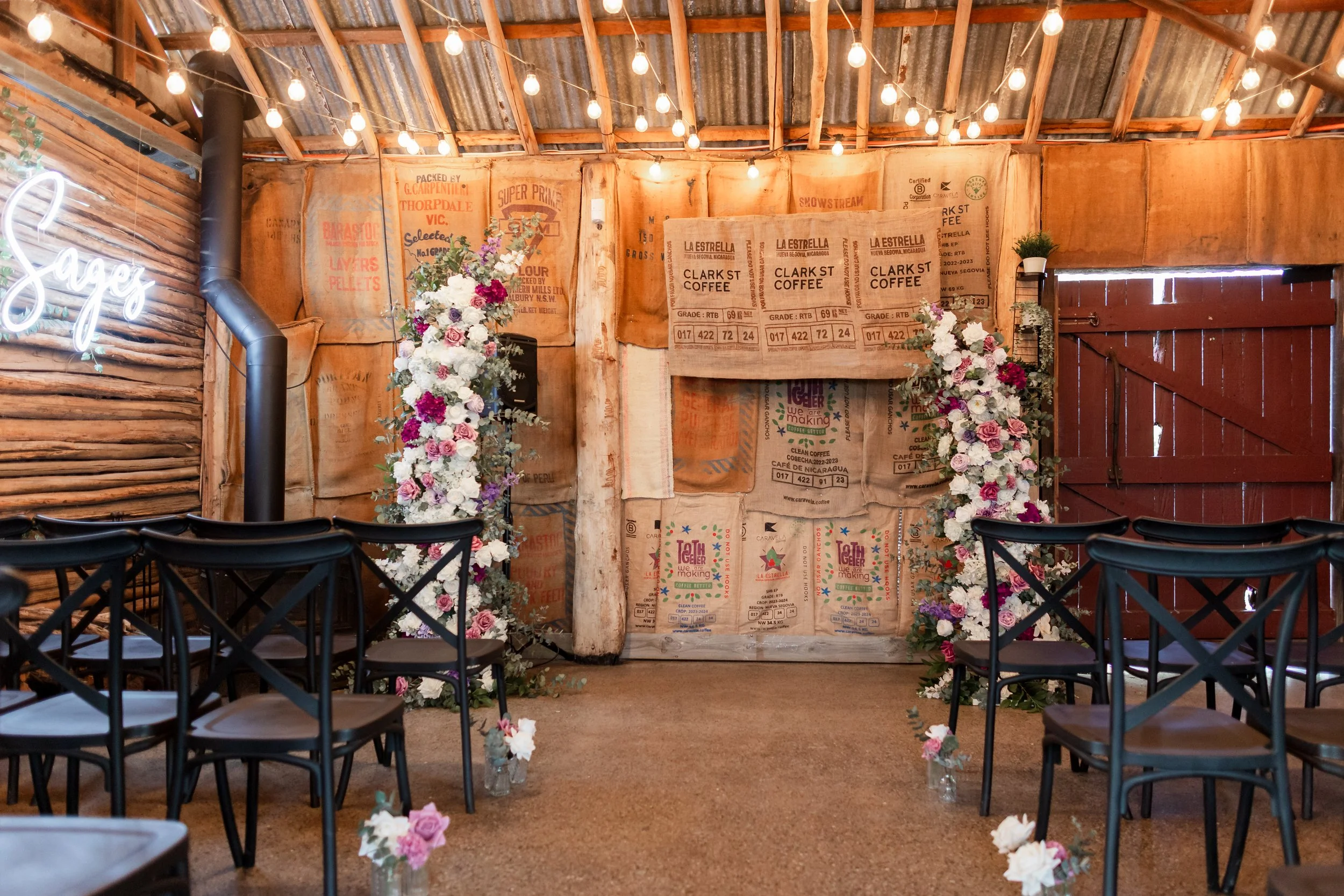 Rustic wedding ceremony setup in a barn with floral arrangements, string lights, chairs, and burlap decorations on the walls.