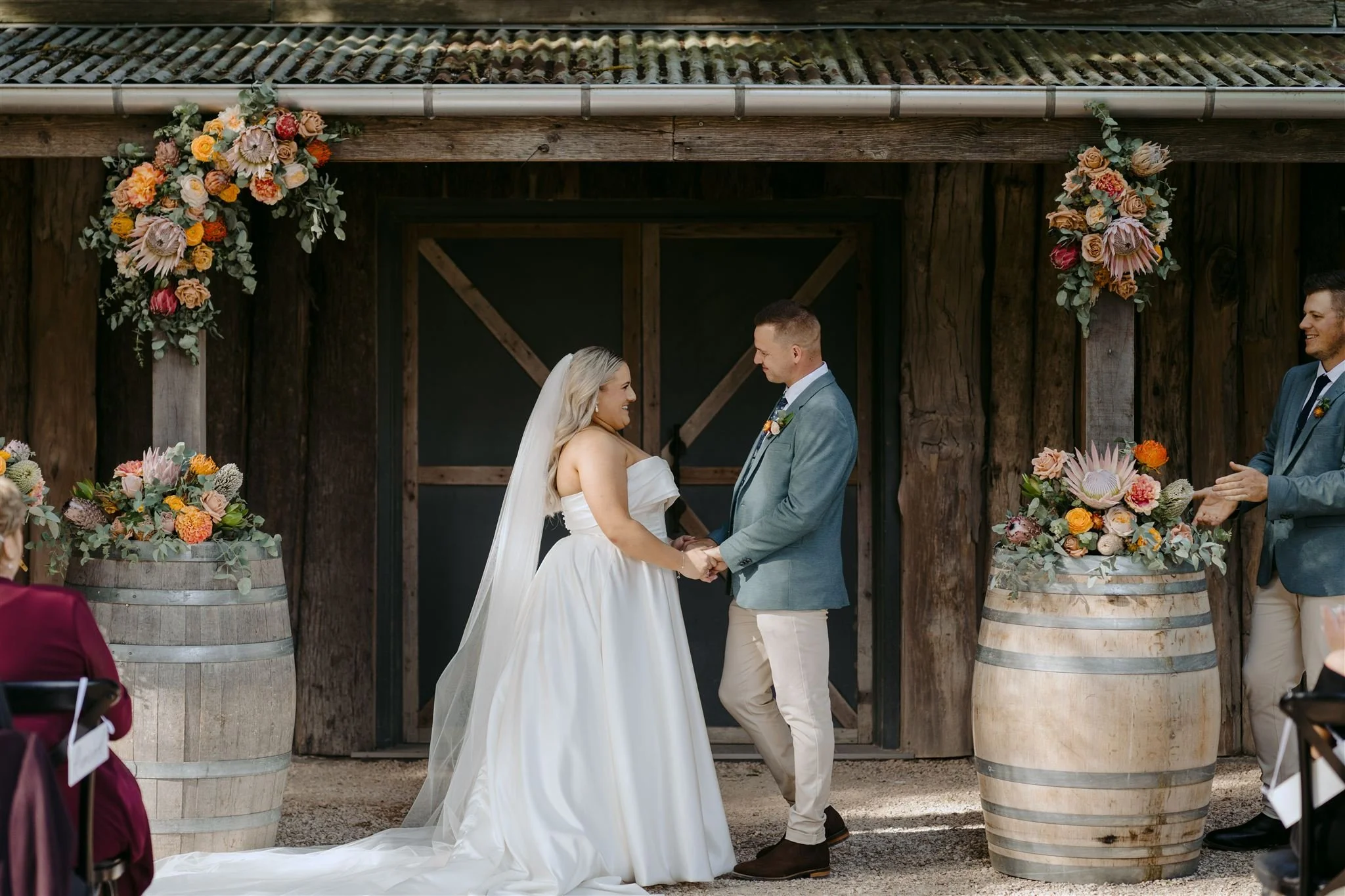 A bride and groom holding hands during their outdoor wedding ceremony, standing in front of a rustic wooden barn decorated with large floral arrangements in barrels, with guests seated nearby.