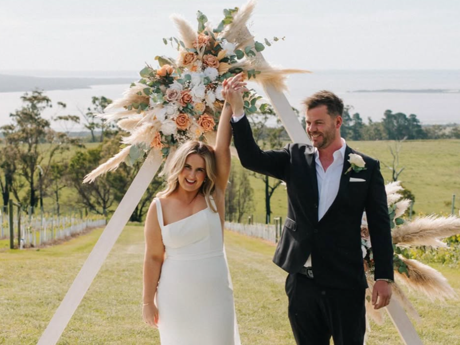A newlywed couple smiling and holding hands in front of a floral wedding arch outdoors with green fields and a body of water in the background.