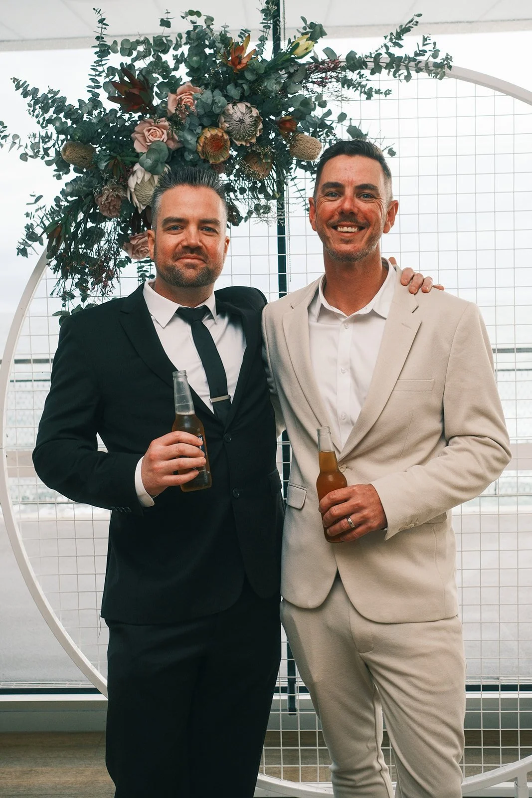 Two men in formal suits holding drinks, standing in front of a large floral arch with greenery and flowers, at a festive event or celebration.