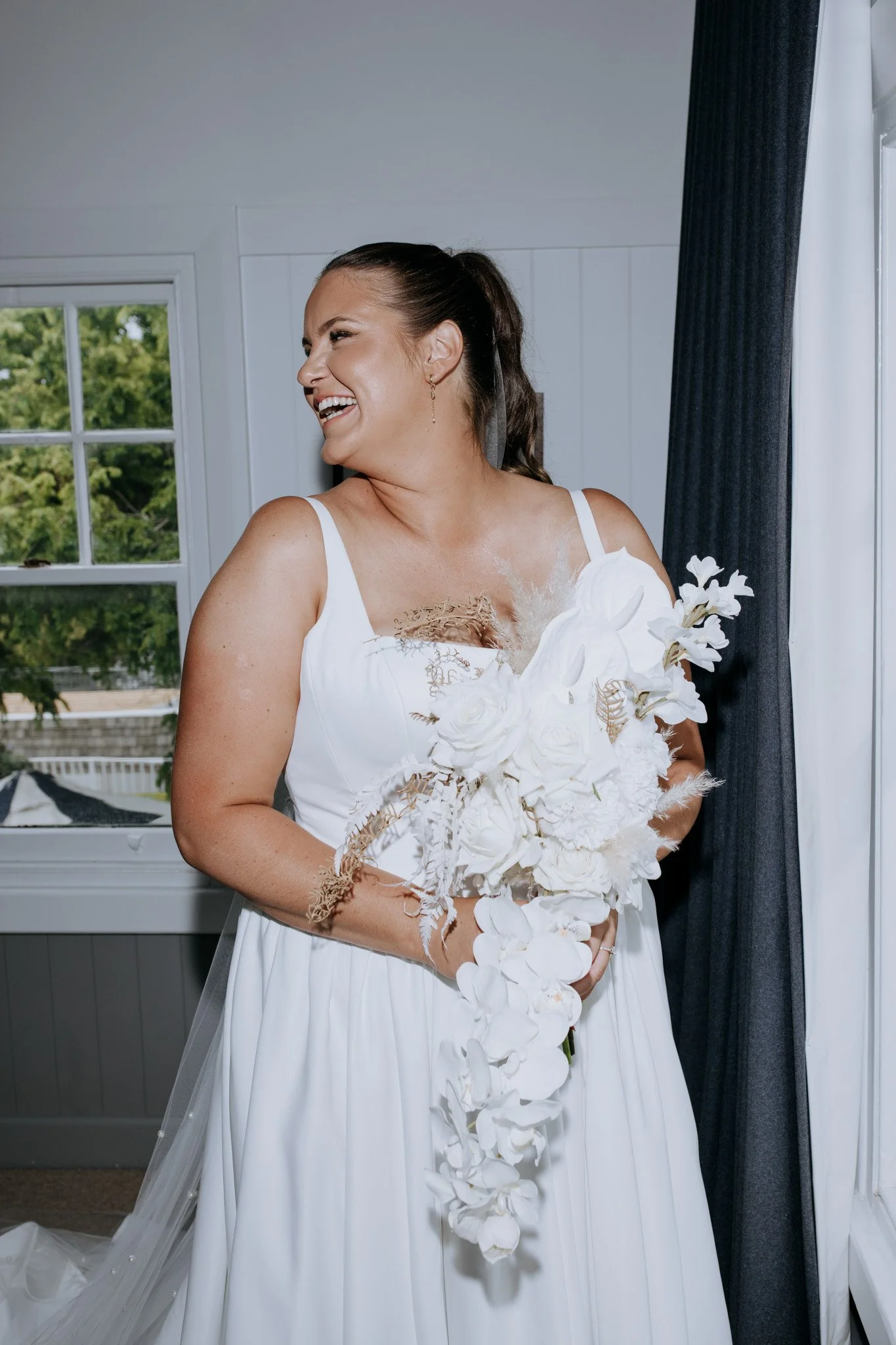 A smiling bride holding a cascading white floral bouquet indoors, with a window showing green trees outside.