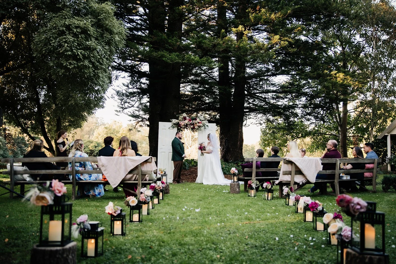 A wedding ceremony taking place outdoors under large trees with hanging branches. The bride and groom stand before a floral arch, exchanging vows. Guests sit on benches on either side, with decorative lanterns and flowers lining the aisle.