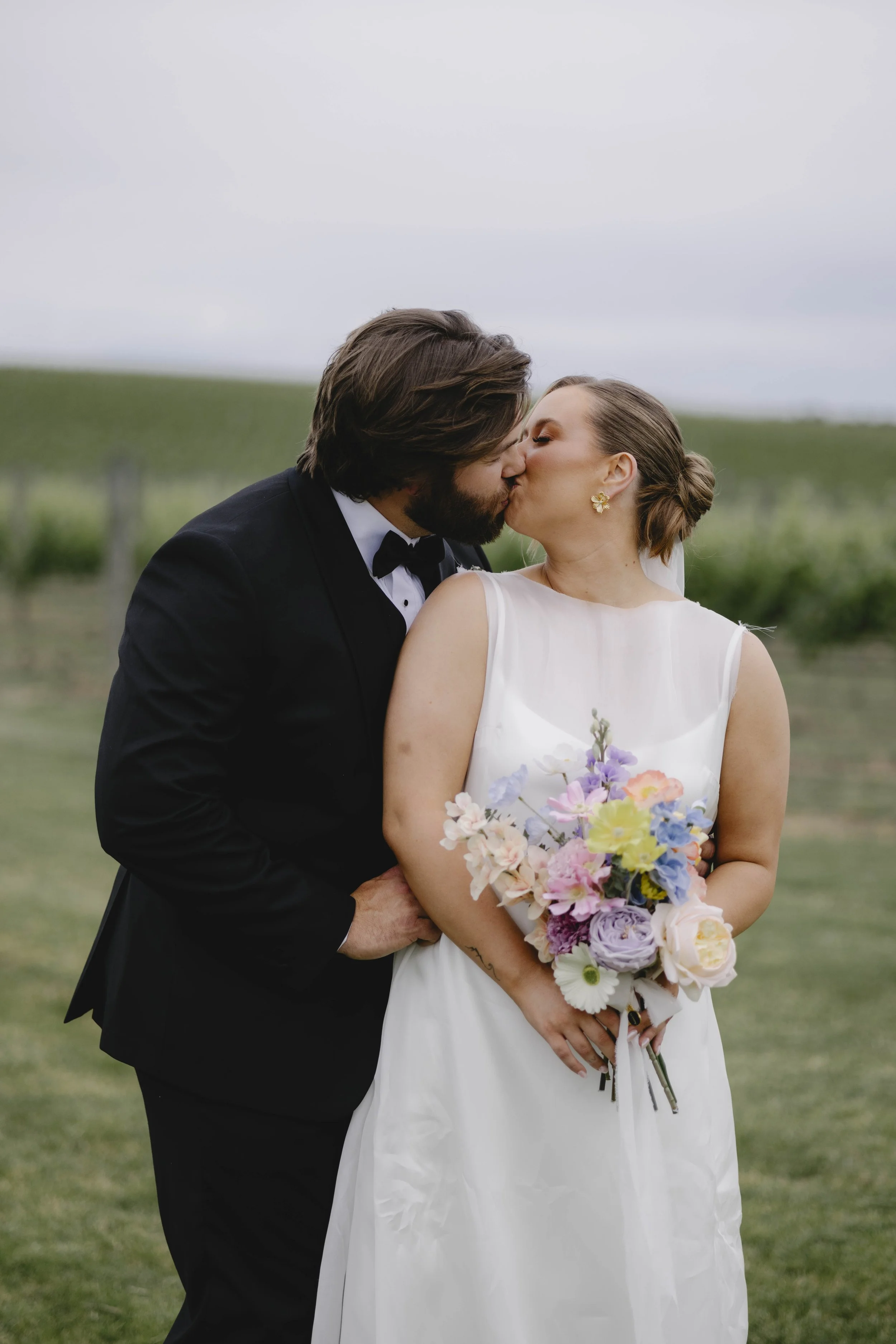 A bride and groom sharing a kiss outdoors during a wedding ceremony, with the bride holding a colorful bouquet of flowers and the groom in a black tuxedo.