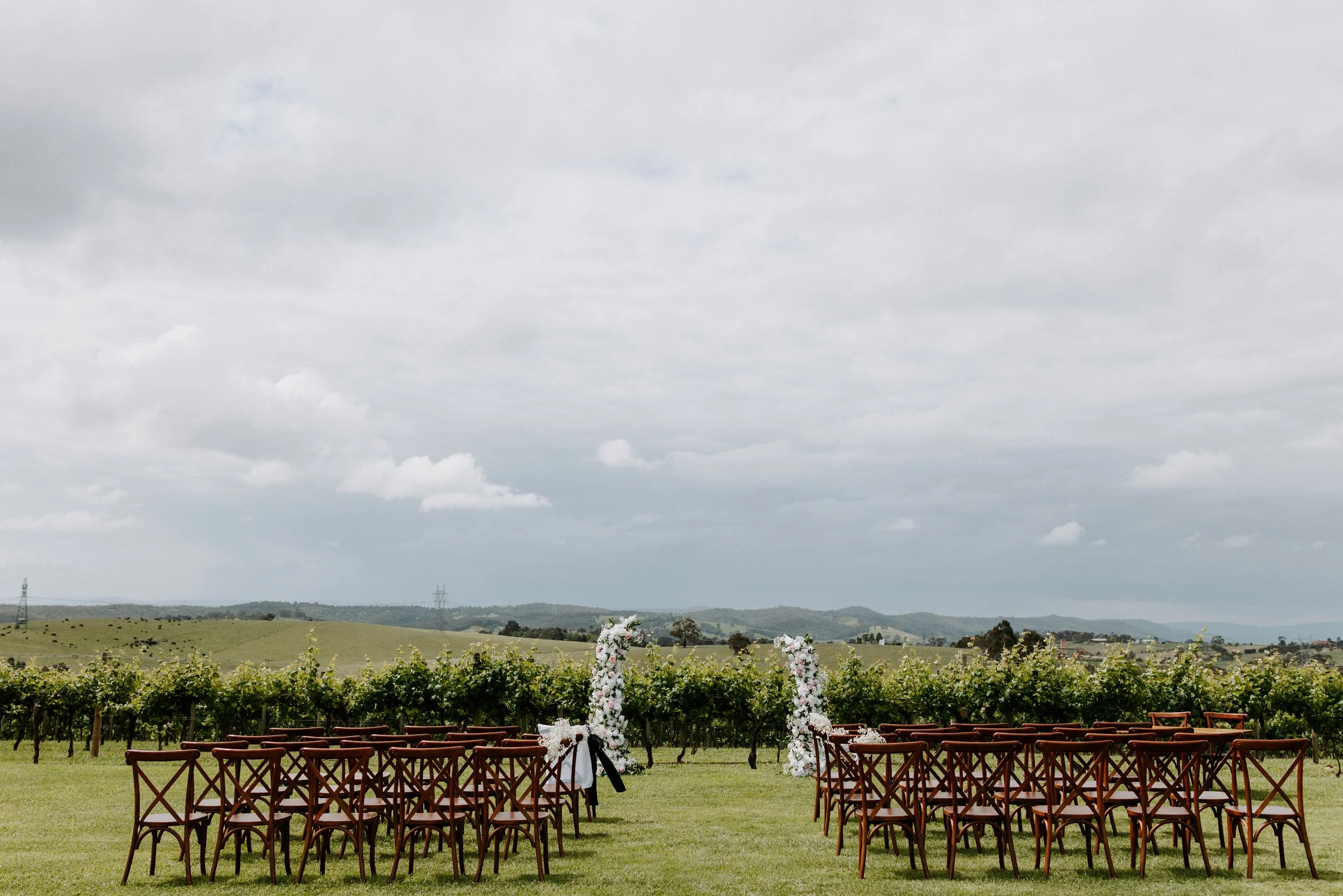 Outdoor wedding setup in a vineyard with rows of chairs facing a floral arch under a cloudy sky.
