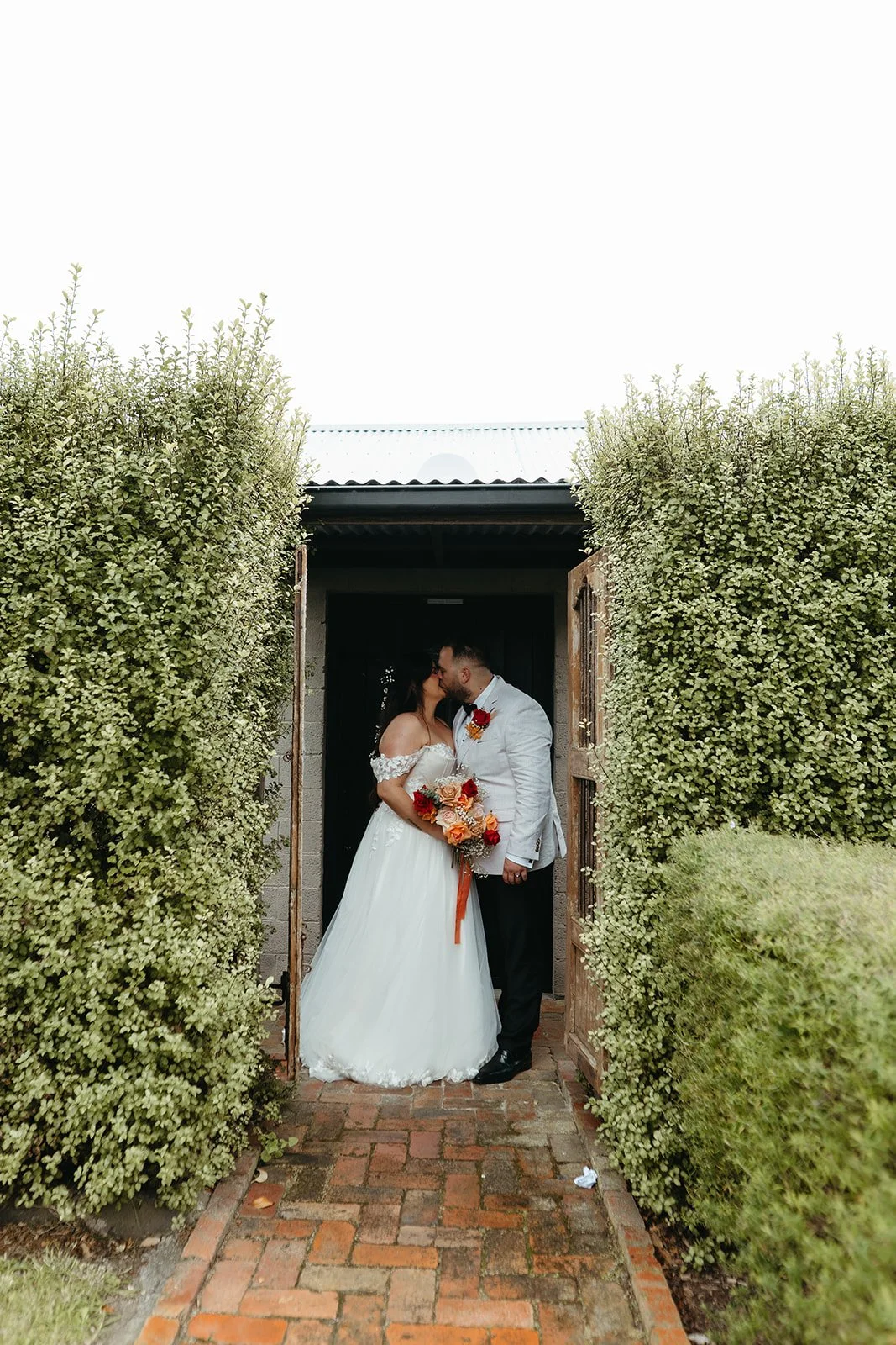 A bride and groom share a kiss in a garden entrance framed by tall green hedges, with the bride holding a bouquet of orange, pink, and red flowers.