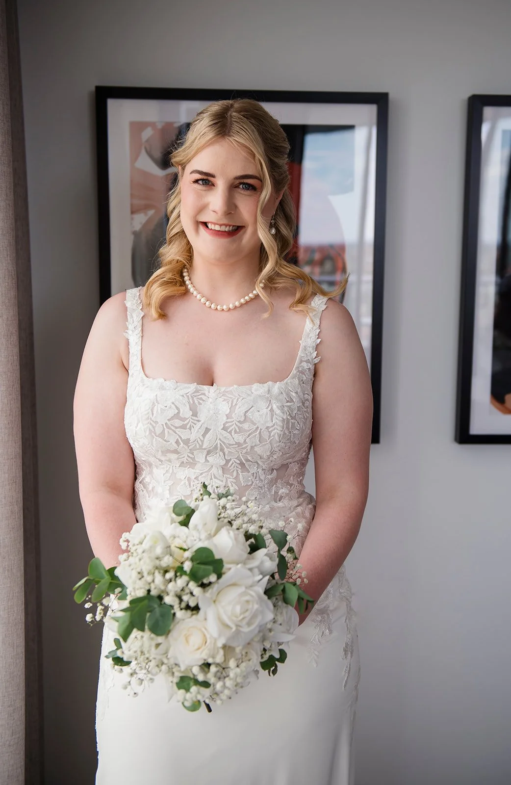 A smiling bride in a white lace wedding dress with a pearl necklace, holding a bouquet of white roses and greenery, standing indoors in front of framed artwork.