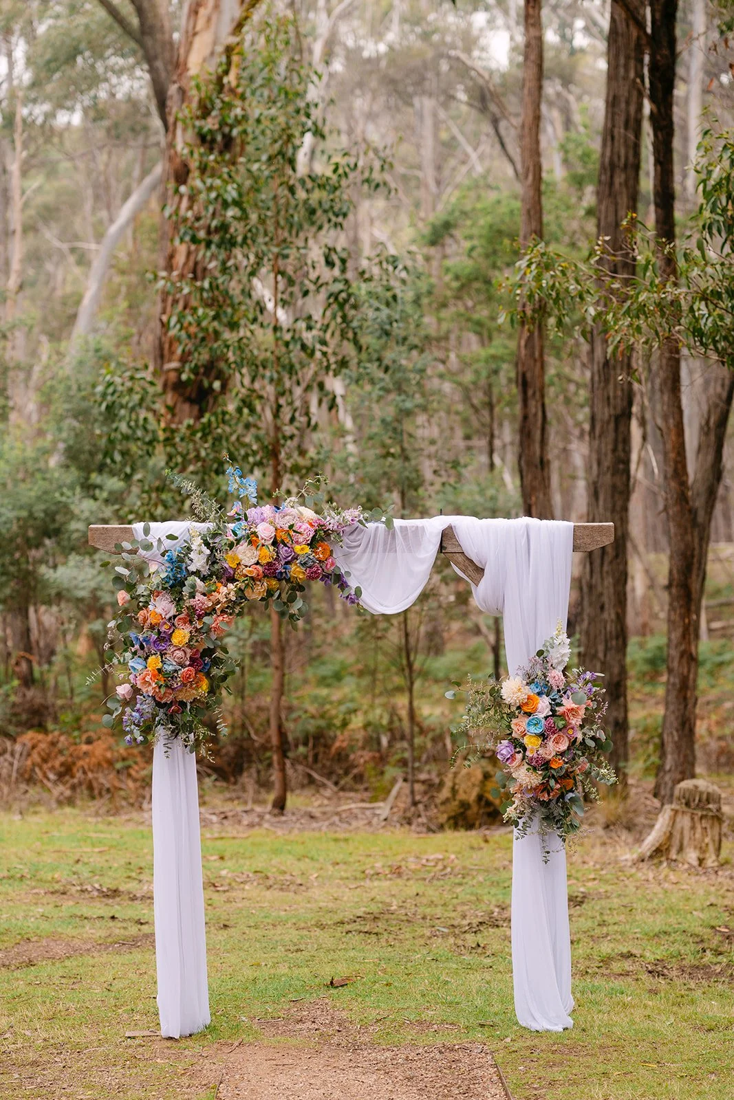 Wedding arch decorated with colorful flowers and white fabric in a forest setting.