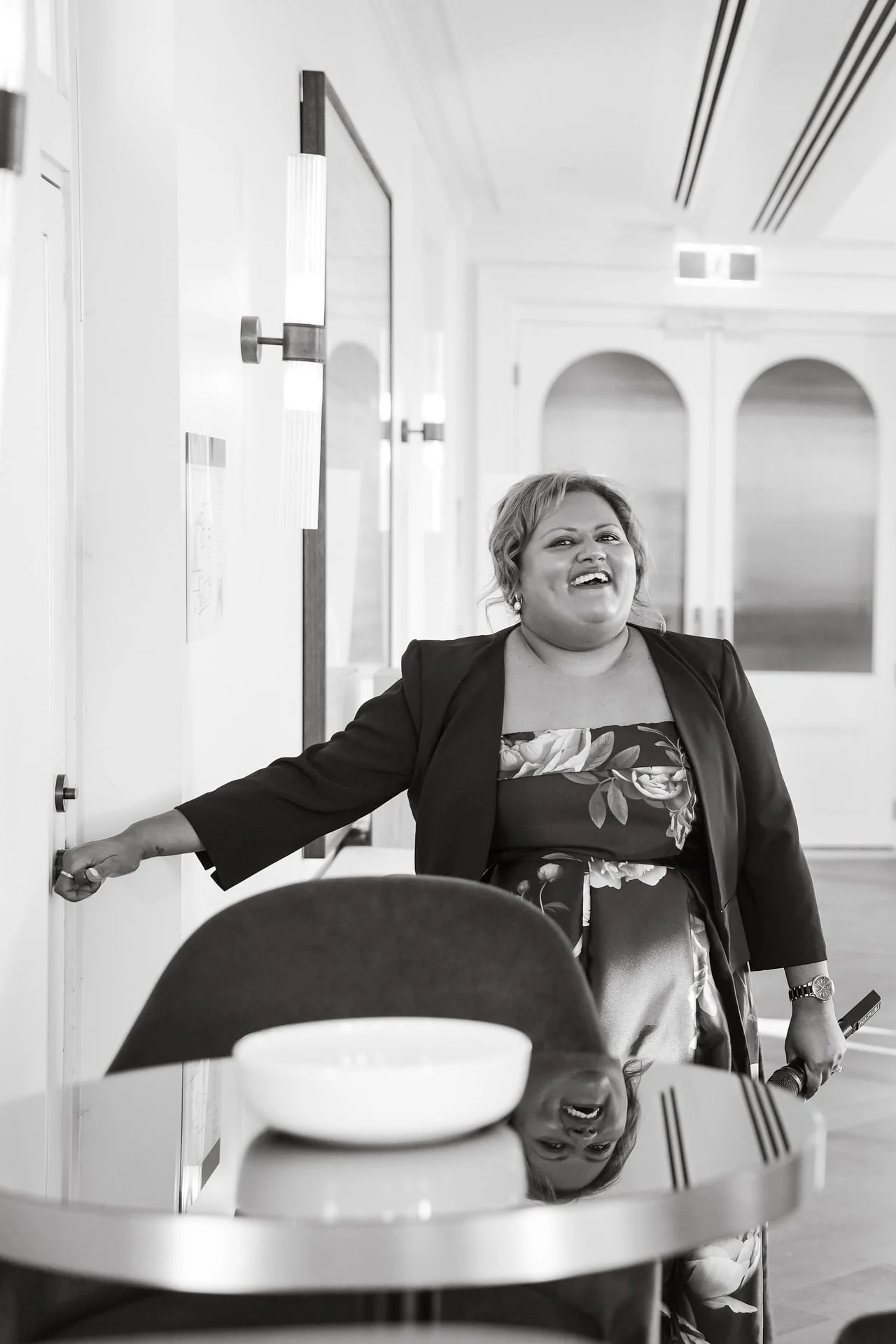 A woman smiling and reaching out with one hand in an indoor setting, with a reflection of her face visible on a shiny surface below a table.