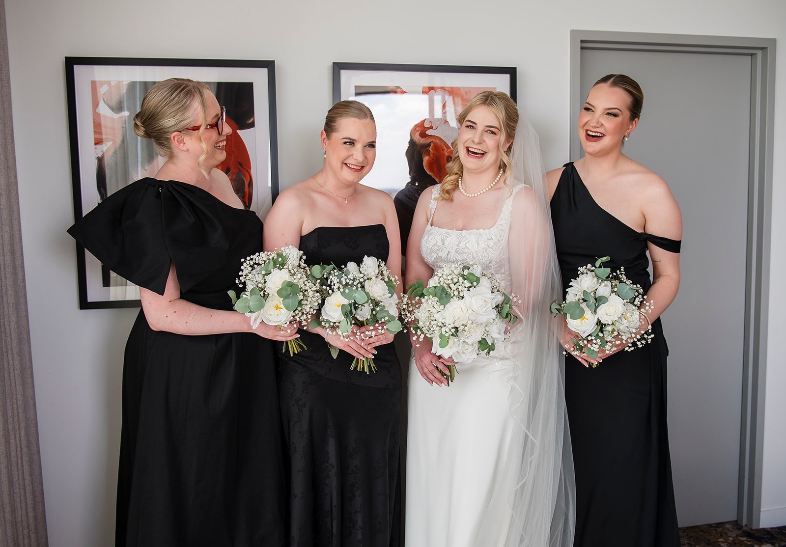 A bride in a white wedding dress with her bridesmaids dressed in black, standing indoors with framed artwork on the wall behind them, all holding bouquets of white flowers.