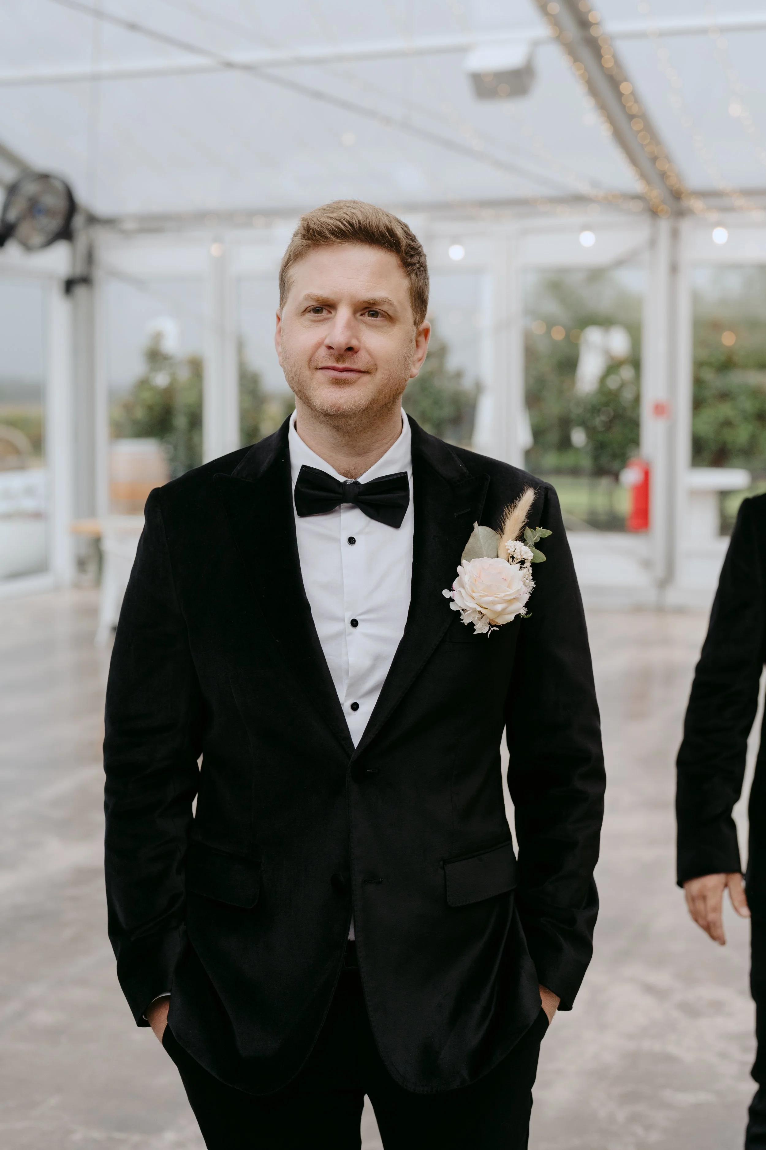 A man in a tuxedo with a bow tie and a boutonnière, standing inside a glass event tent with string lights, looking into the camera.