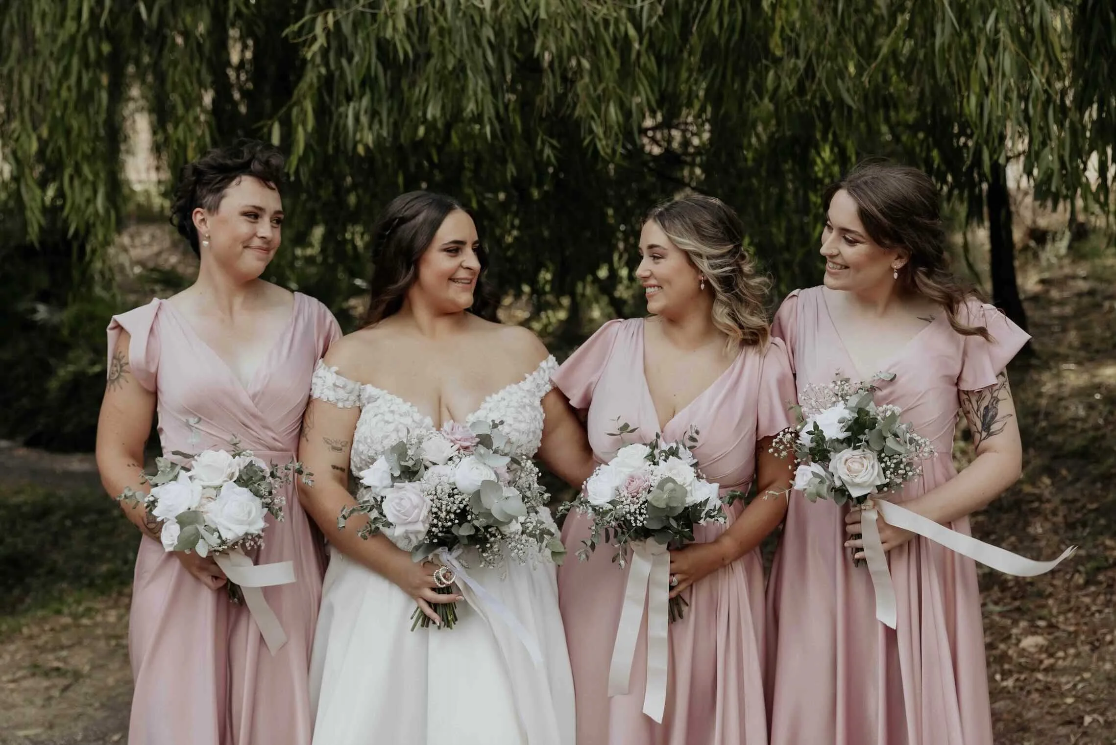 Bride in off-shoulder gown with bridesmaids in dusty pink, all holding white and blush rose bouquets with trailing ribbon