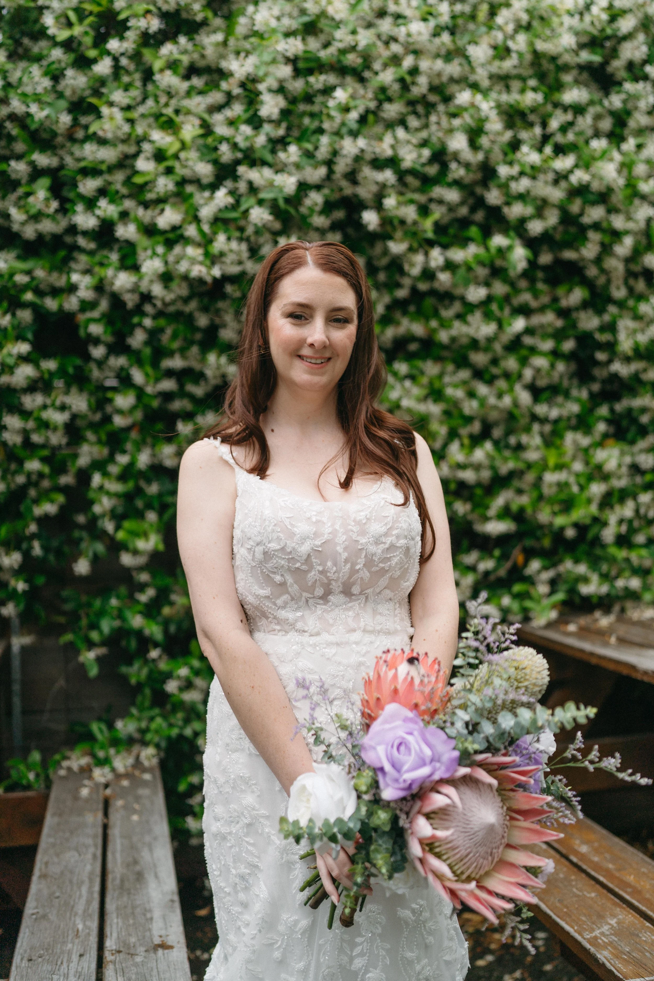 A woman in a white wedding dress holding a bouquet of flowers, standing outdoors in front of a lush, flowering green hedge.