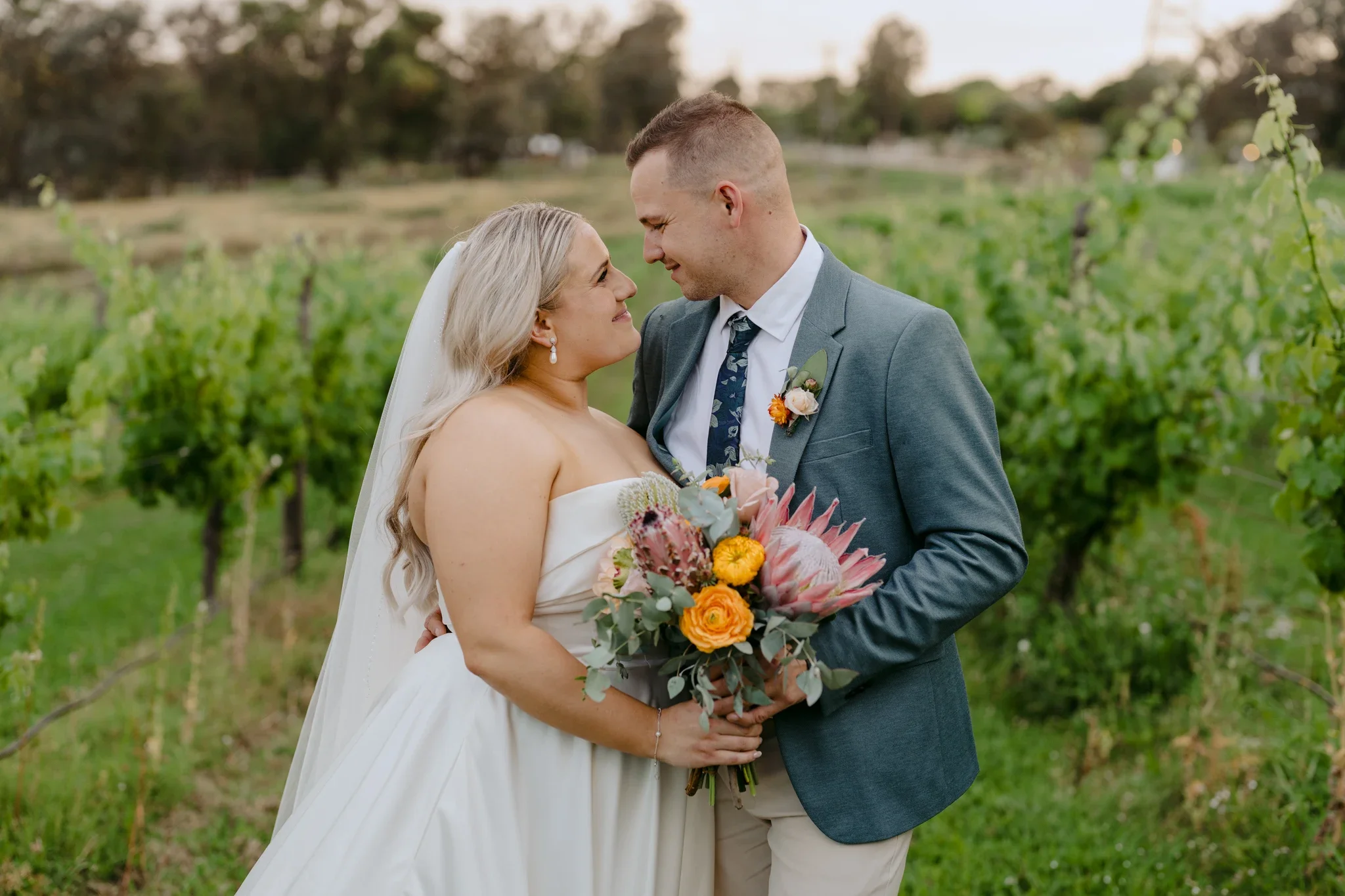A bride and groom standing close together holding a bouquet of flowers outdoors in a vineyard during sunset.