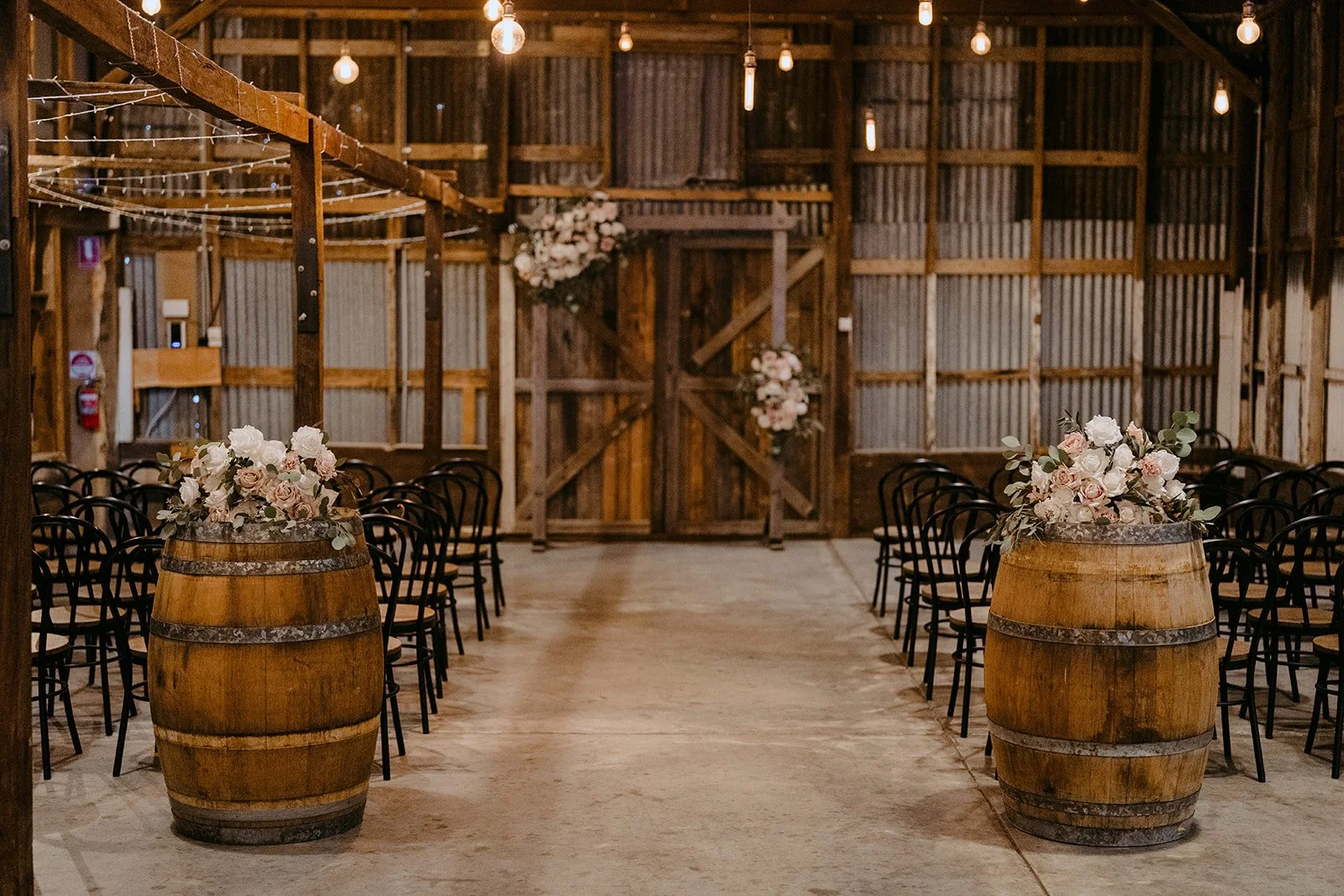 Rustic wedding ceremony setup with wooden barrels topped with floral arrangements, black chairs arranged on either side, and a wooden barn interior decorated with flowers and string lights.