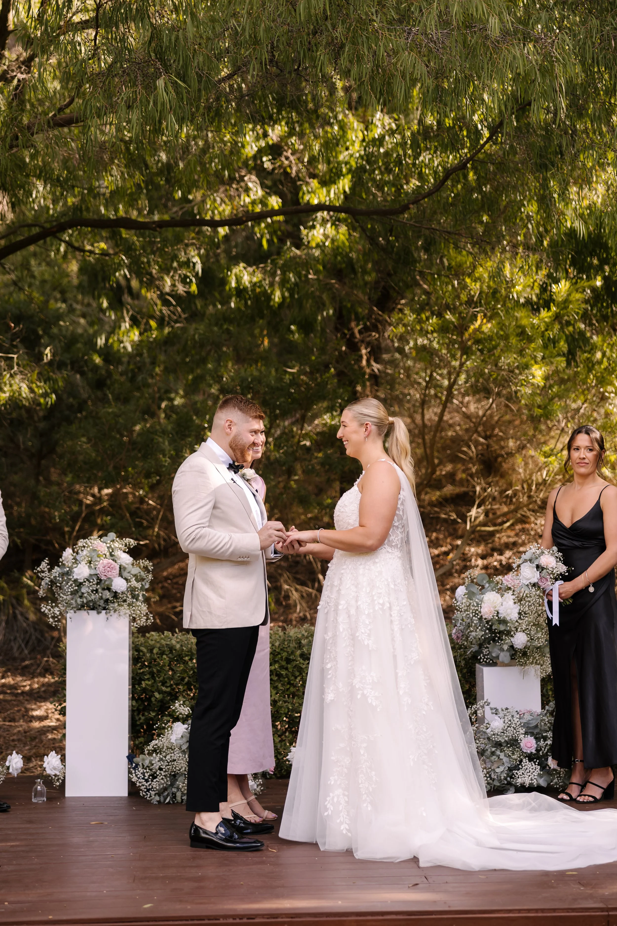 A wedding ceremony outdoors with a bride and groom exchanging rings, flanked by bridesmaids and floral arrangements on a wooden platform under trees.
