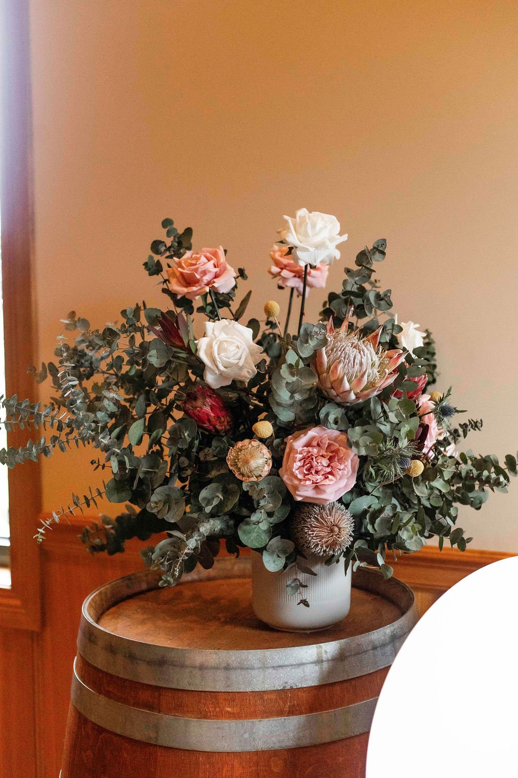A bouquet of mixed flowers including roses, protea, and other blooms with eucalyptus leaves in a white vase on a wooden barrel.