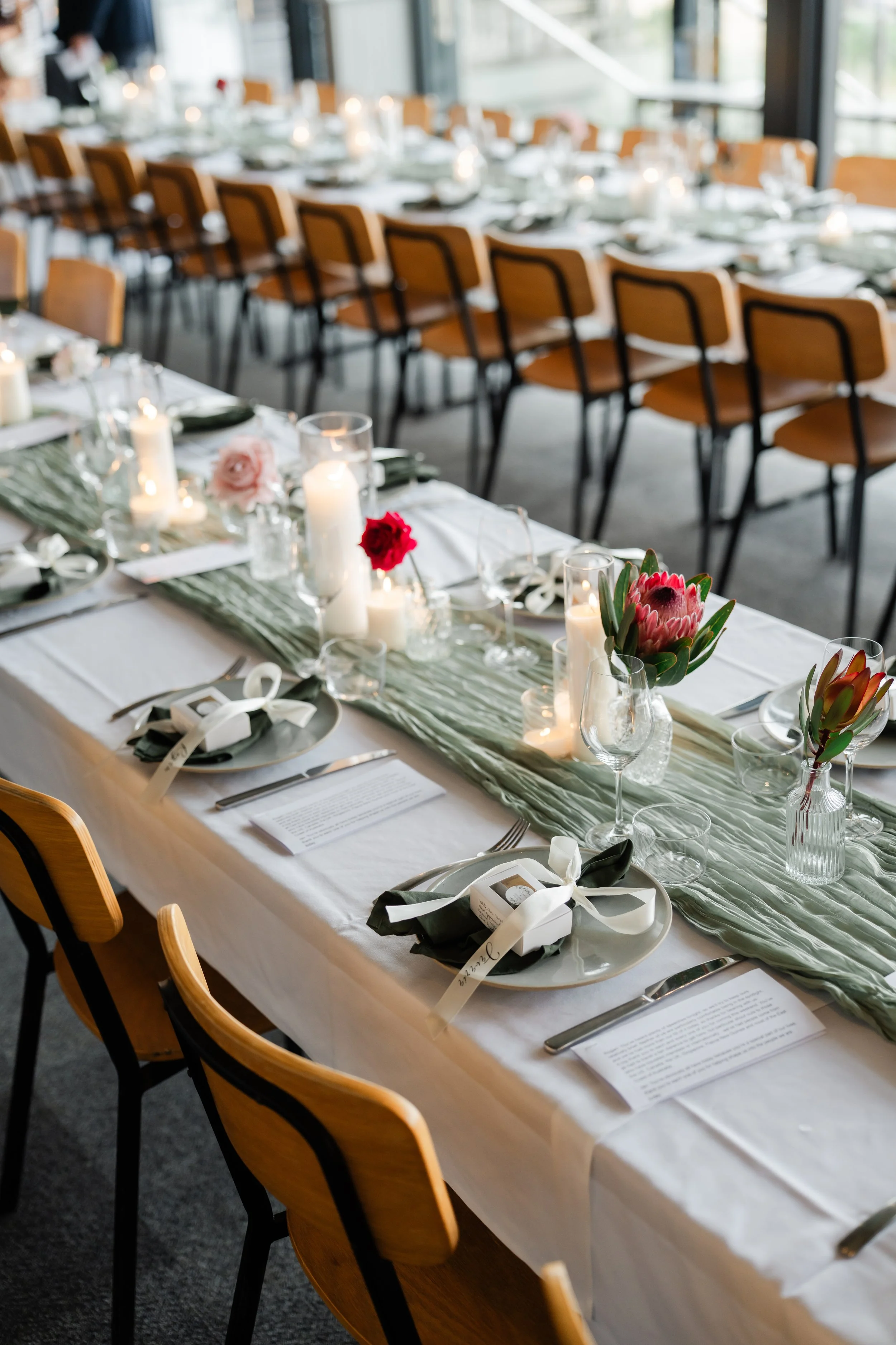A white rose with gum leaves and babys breath in an amber coloured vase sits on a wedding table surrounded by plates and candles