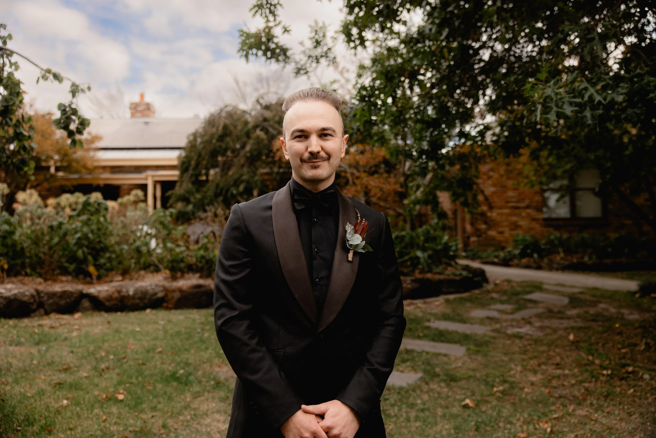 A man in a black tuxedo with a boutonniere stands outdoors in front of a garden and house, smiling at the camera during daytime.