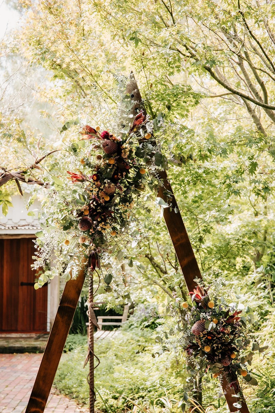 A decorated wooden arch with plentiful floral arrangements, set outdoors among green trees and a garden.