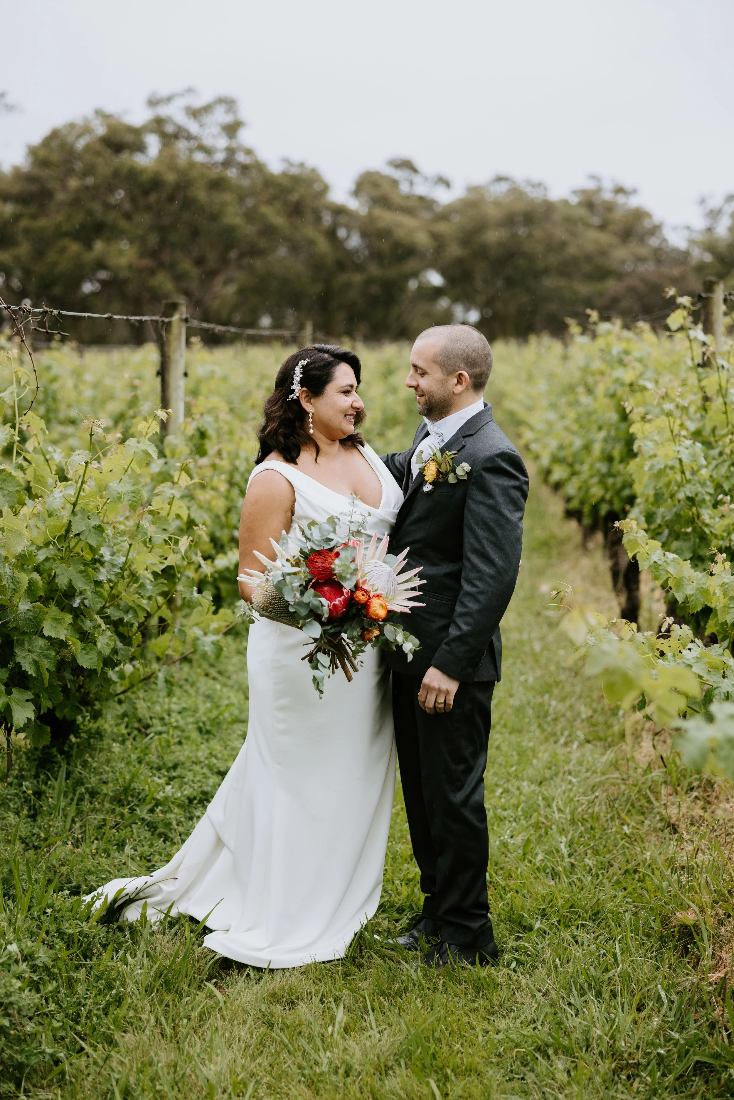 A bride and groom standing in a vineyard, smiling at each other, with the bride holding a bouquet of flowers.