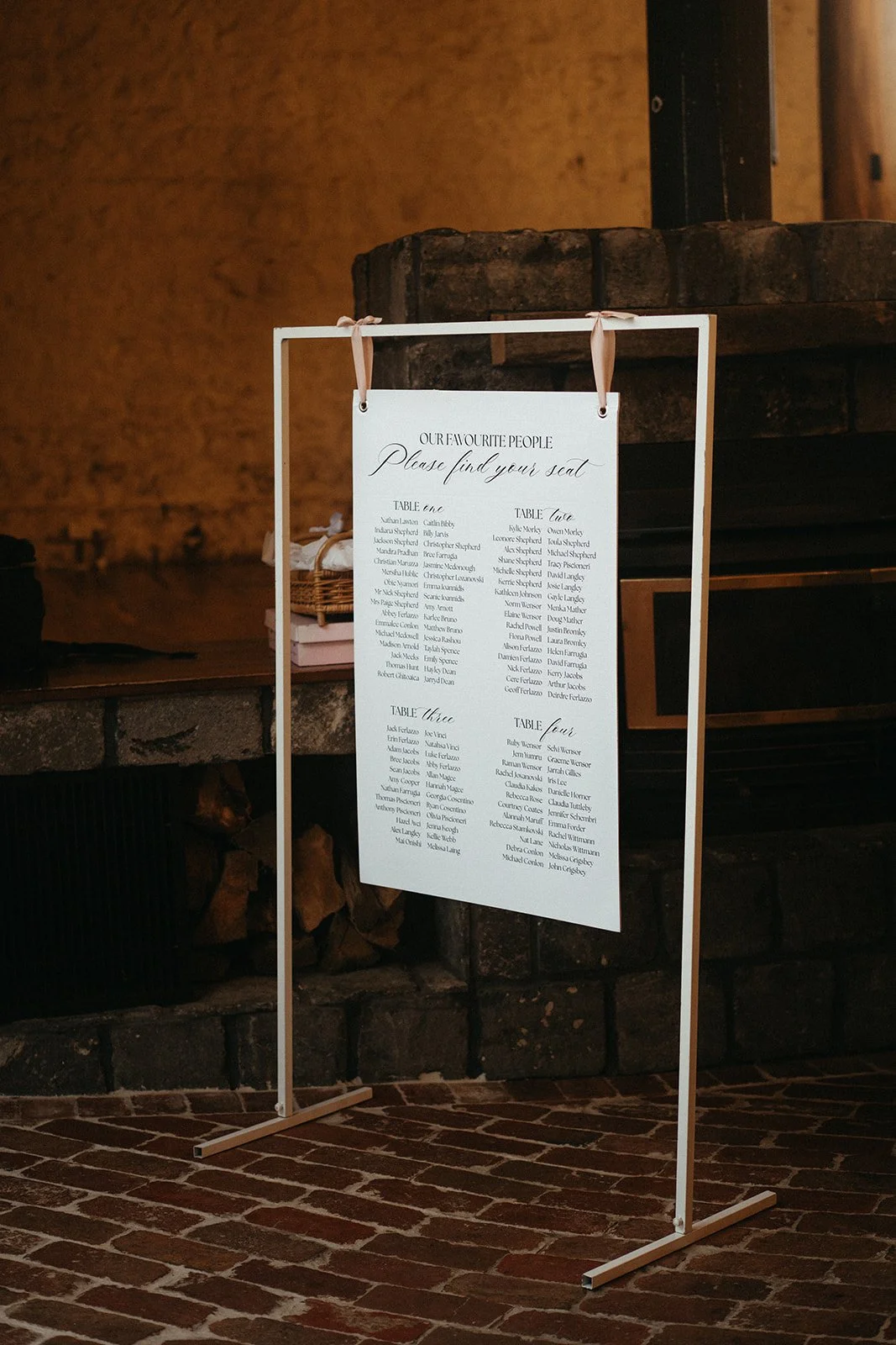 A wedding seating chart on a white stand, hanging with peach ribbons. The chart lists tables and guest names, with a brick fireplace in the background.