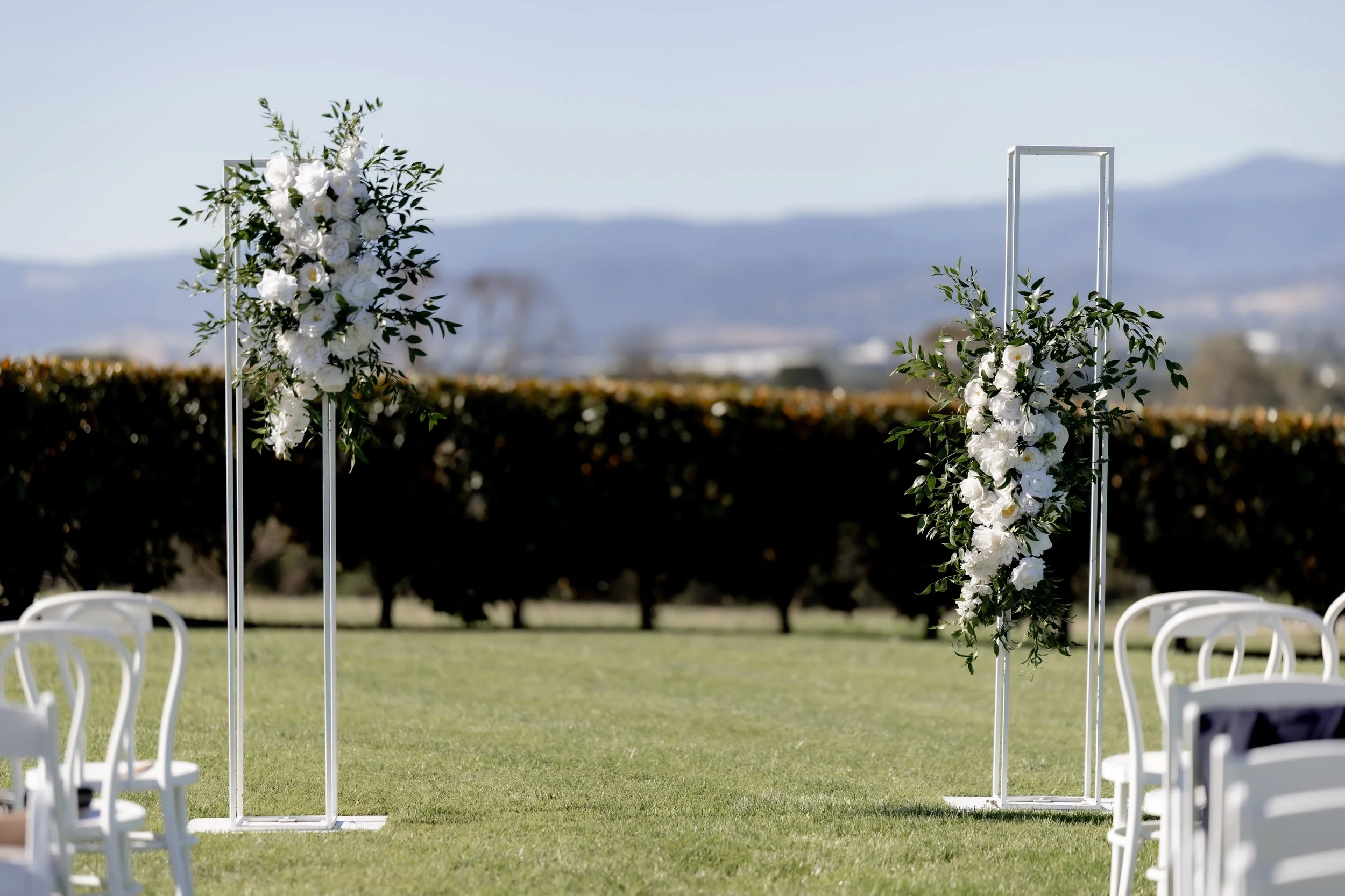 Wedding ceremony setup outdoors with white chairs and floral arch decorations on a grassy field, with mountains in the background.