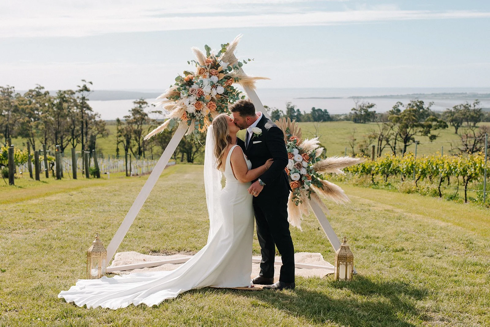A bride and groom kissing under a wedding arch decorated with flowers and greenery outdoors, with a rural landscape and vineyard in the background.