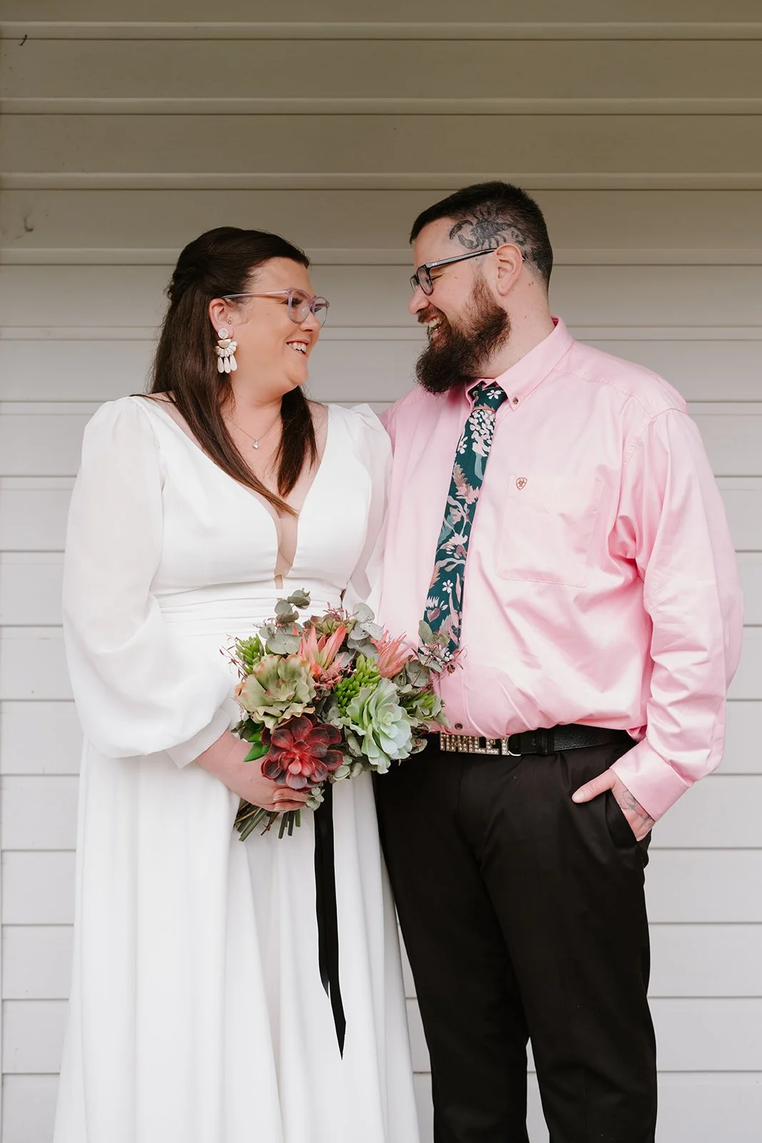 Couple smiling and looking at each other, holding a bouquet, standing against a light-colored wooden wall.