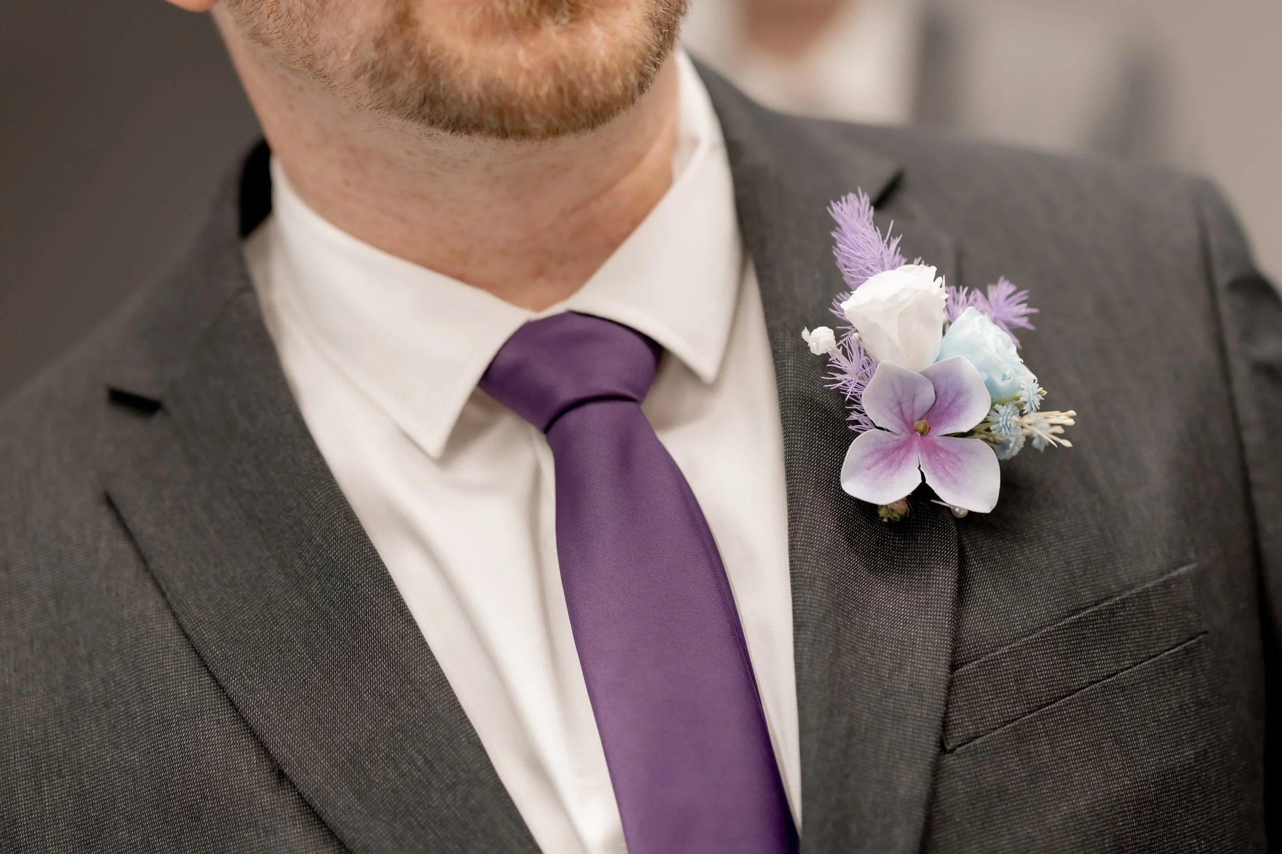 Close-up of a man in a gray suit with a purple tie, showcasing a boutonniere with purple and white flowers on his lapel.