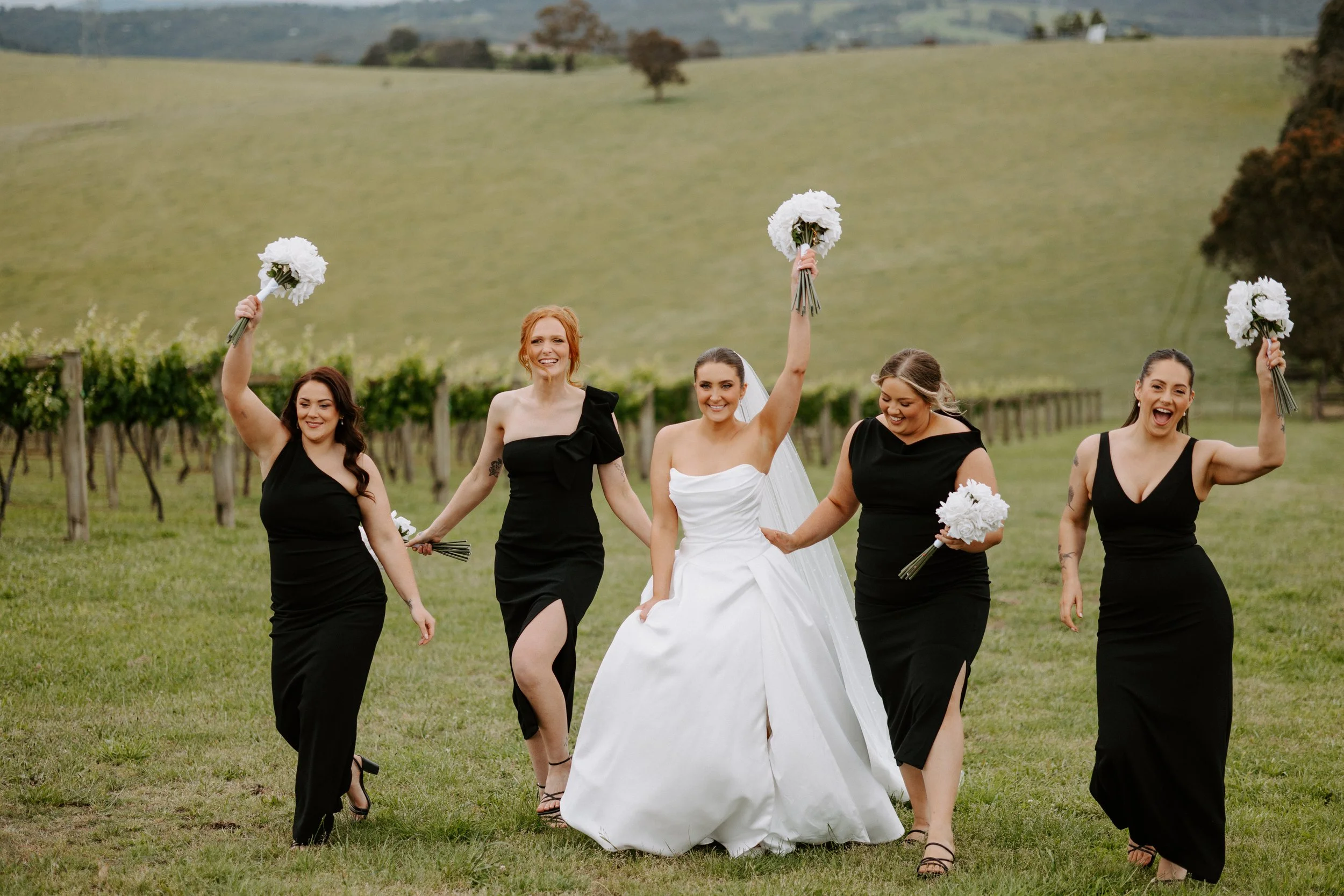 A bride in a white wedding dress and four bridesmaids in black dresses celebrating outdoors in a vineyard, holding bouquets of white flowers and walking across a grassy field.