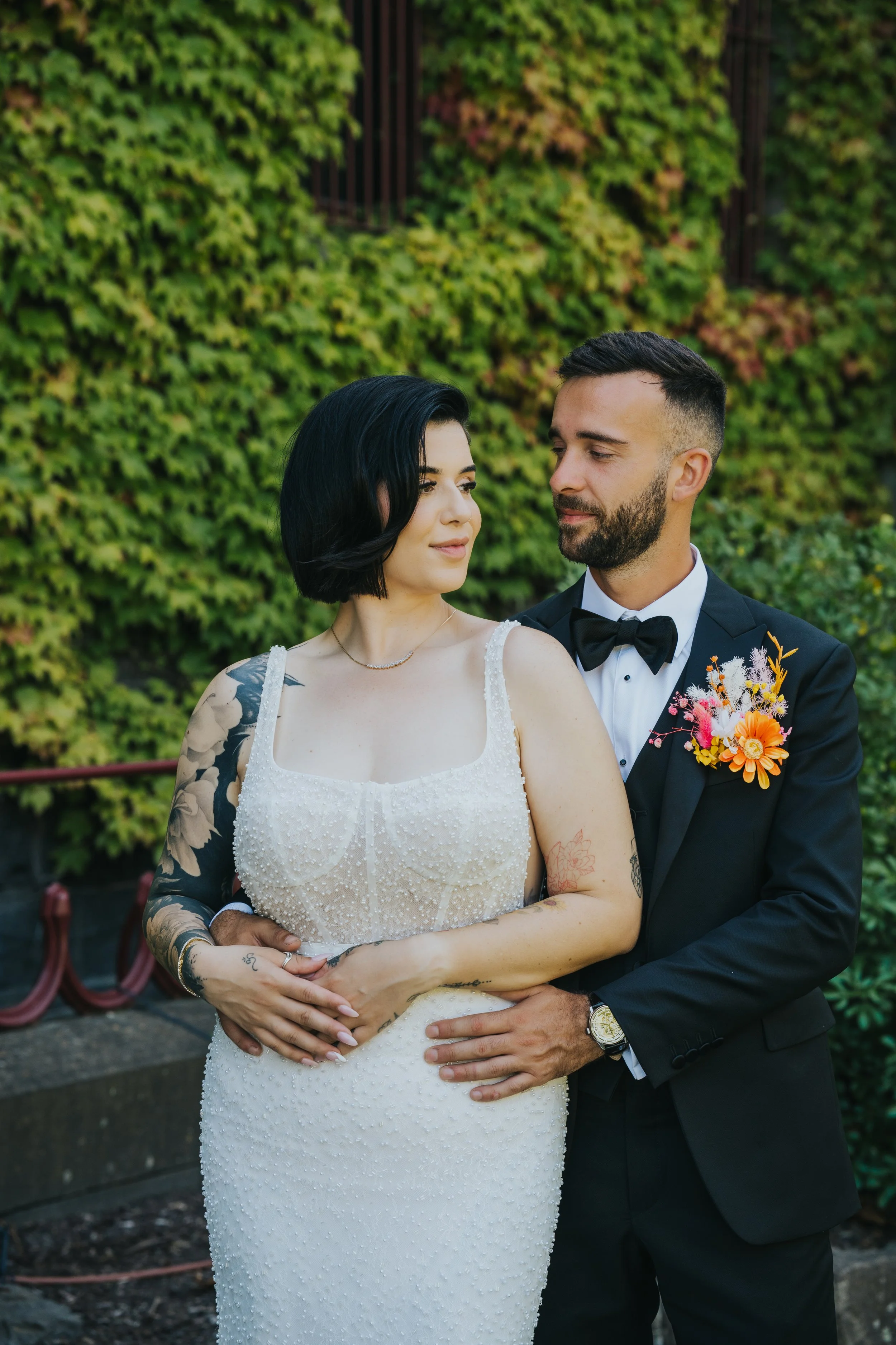 A bride and groom in wedding attire standing closely outdoors, with green foliage in the background. The bride has dark hair, tattoos on her arm, and is wearing a white beaded wedding dress. The groom has short dark hair, a beard, and is dressed in a