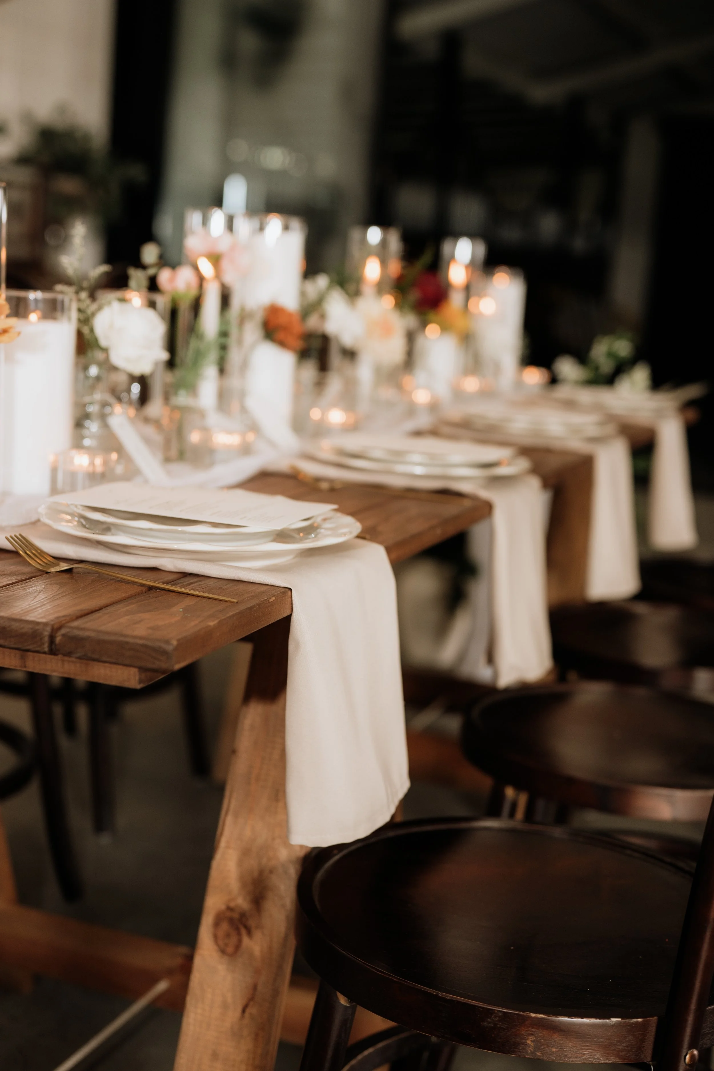 A rustic dining table set with plates, glasses of water, and candles in glass holders, decorated with flowers. The table has a white cloth draped over it and is surrounded by dark wooden chairs. The background shows a dimly lit ambiance, possibly in a barn or a warehouse-style venue.
