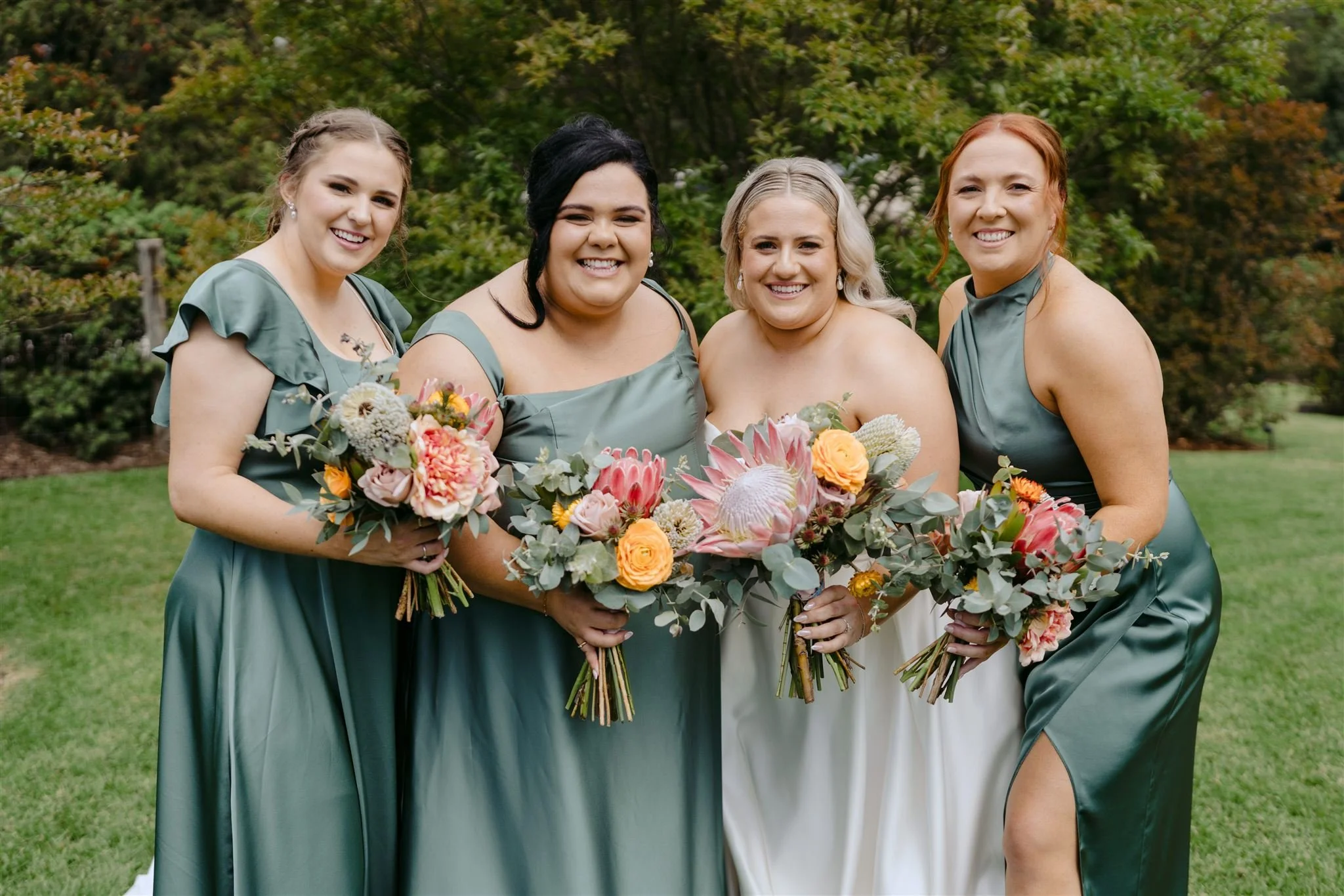 Four women in green and white dresses holding bouquets of flowers, standing outdoors on a grassy area with trees in the background.