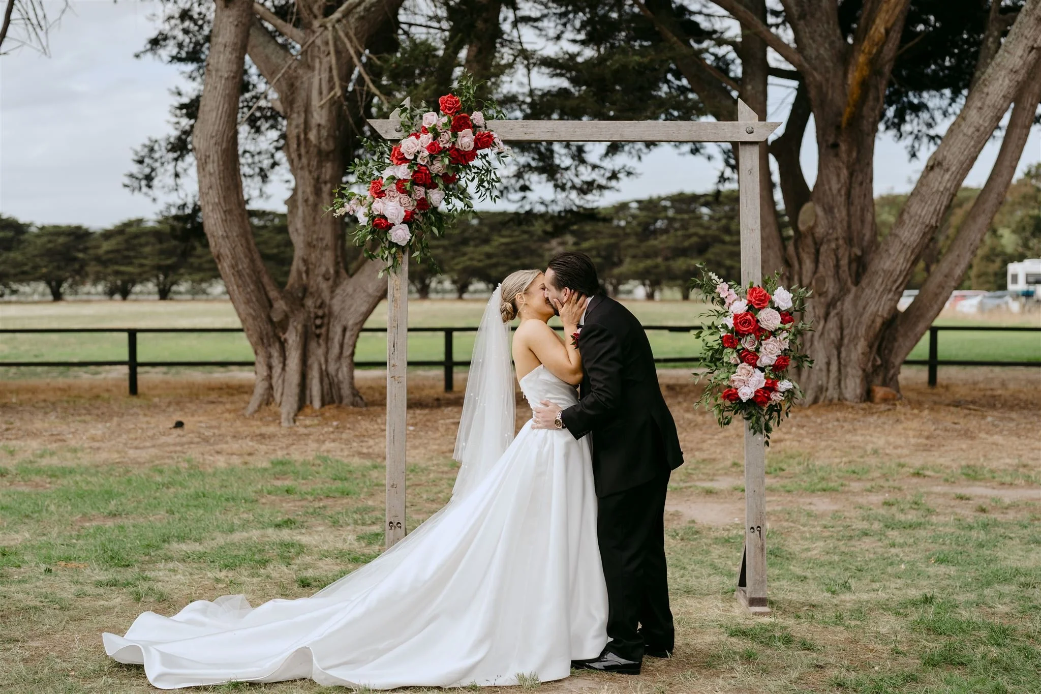 A bride and groom kissing beneath a wooden wedding arch decorated with red and pink roses and greenery on a grassy outdoor setting with trees in the background.