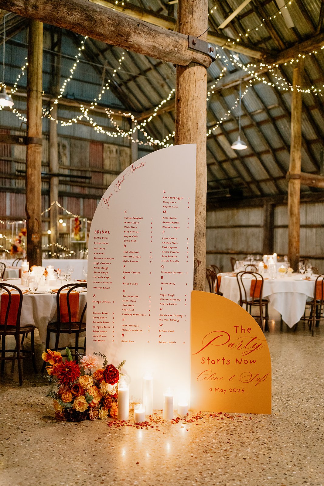 A two-panel wedding seating chart in white and burnt orange, displayed in a fairy-lit rustic barn. Styled with pillar candles and autumn florals in red, orange and peach.