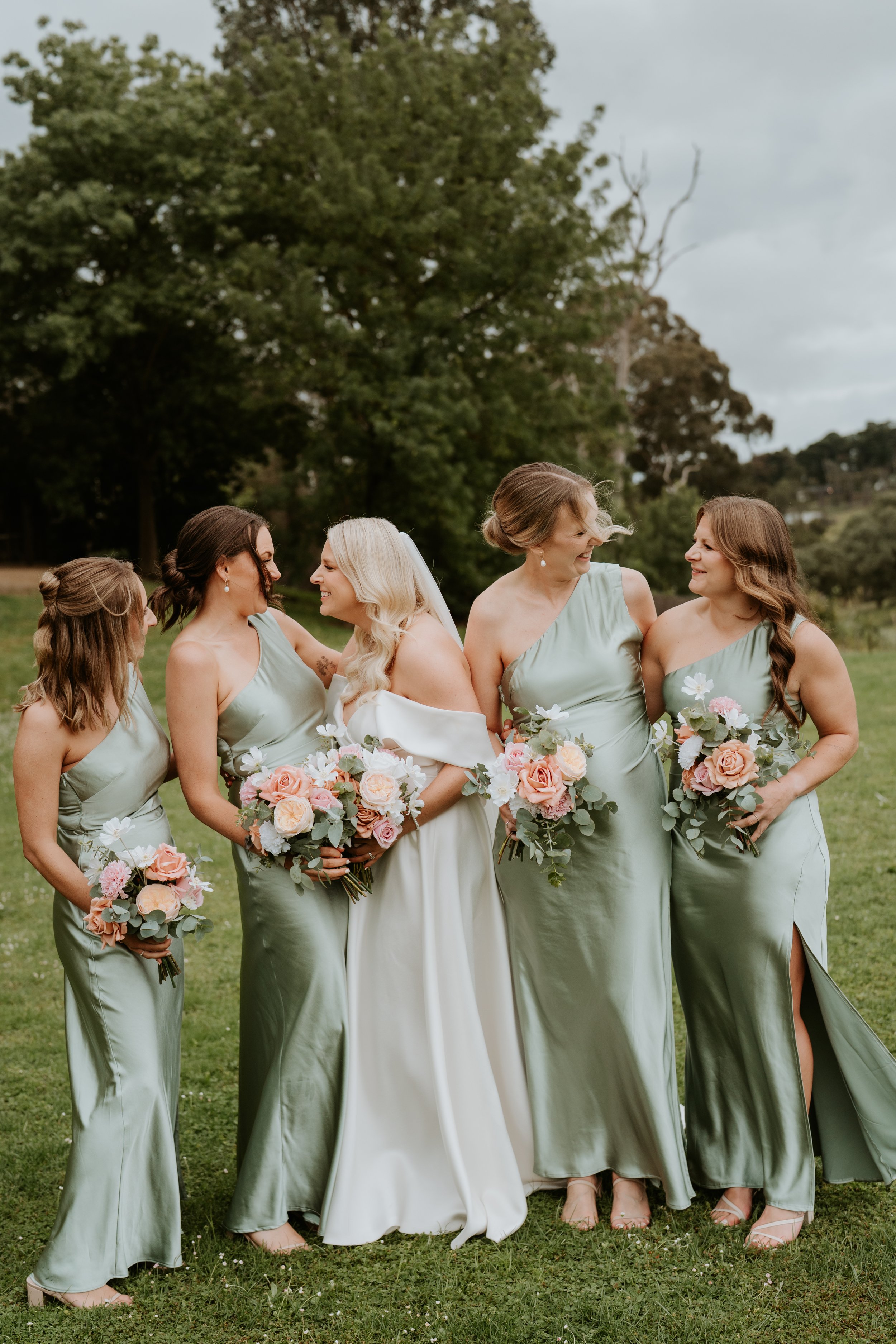 A bride with four bridesmaids outdoors on a grassy area with trees in the background. They are smiling and holding bouquets of pink, peach, and white flowers with greenery. The women are wearing matching sage green dresses.