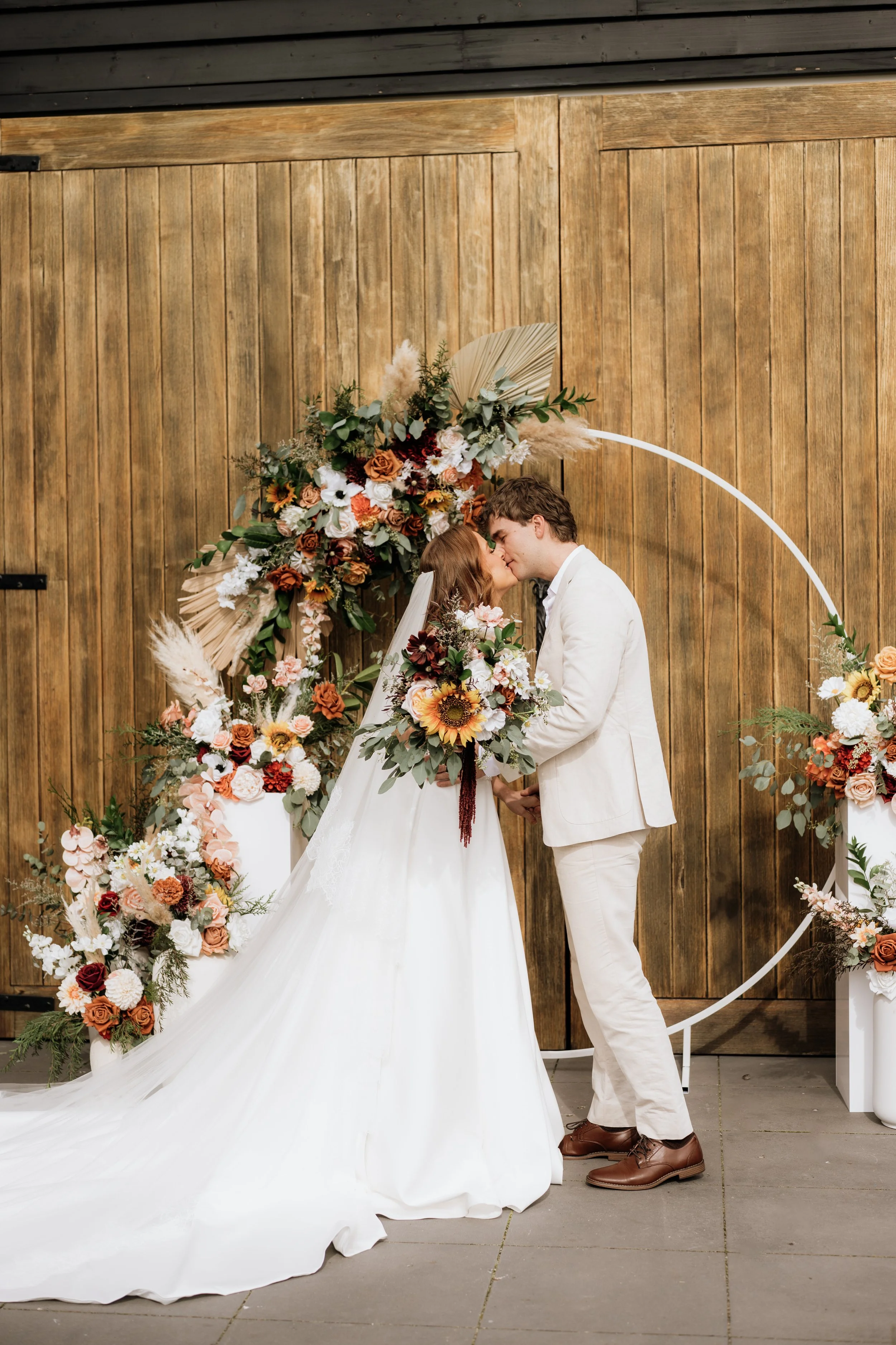A couple in wedding attire sharing a kiss during their wedding ceremony, standing in front of a floral arrangement and wooden background.