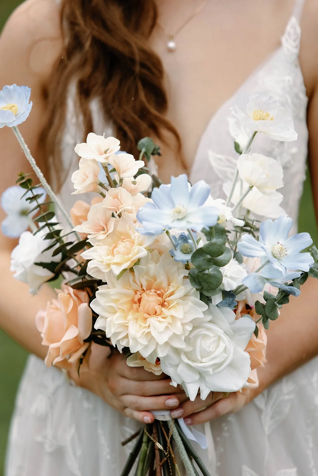 A woman in a wedding dress holding a bouquet of pastel-colored flowers including cream, peach, and light blue blooms.