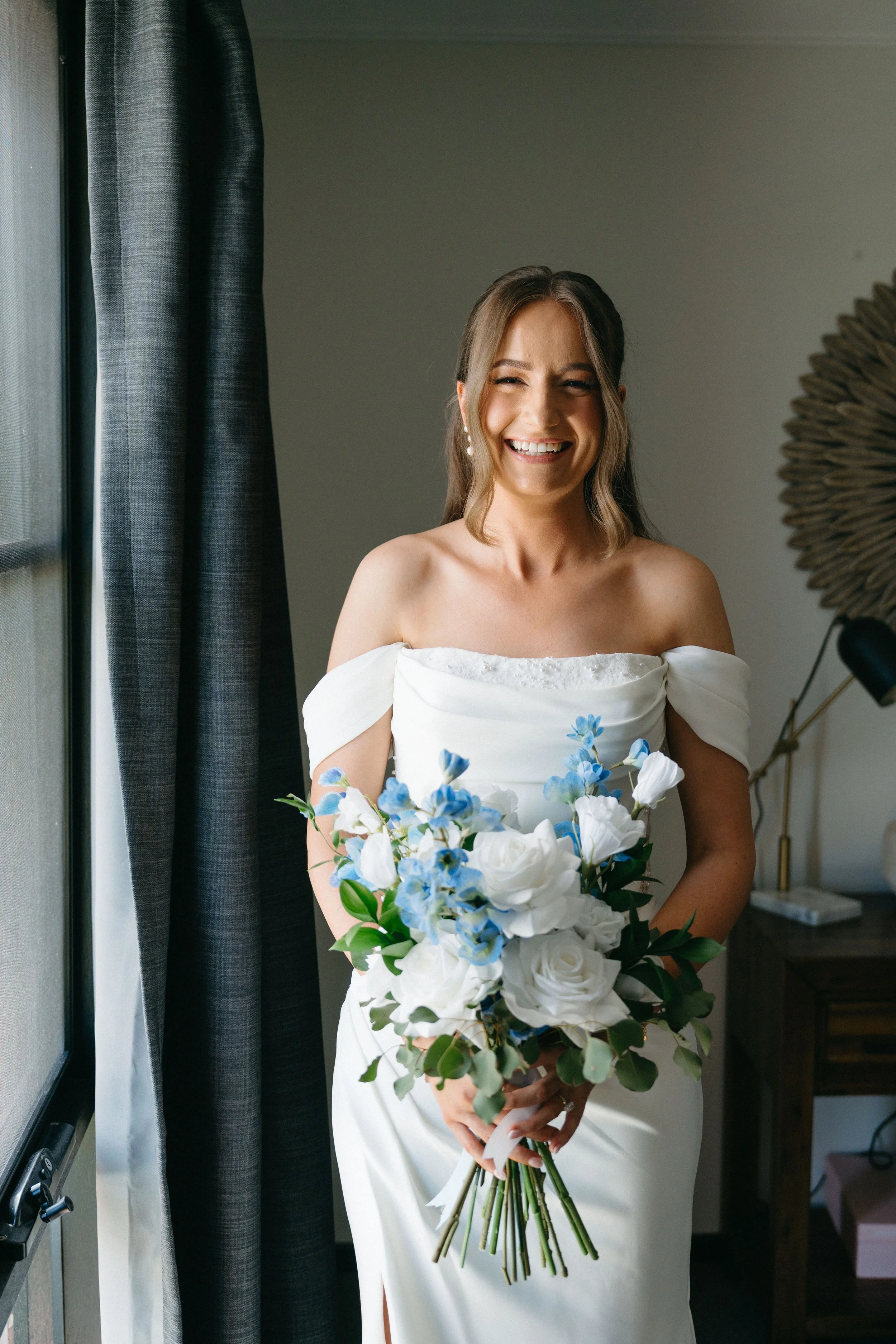 A smiling bride in a white off-shoulder wedding dress holding a bouquet of white and blue flowers, standing next to a window with dark curtains in a room.