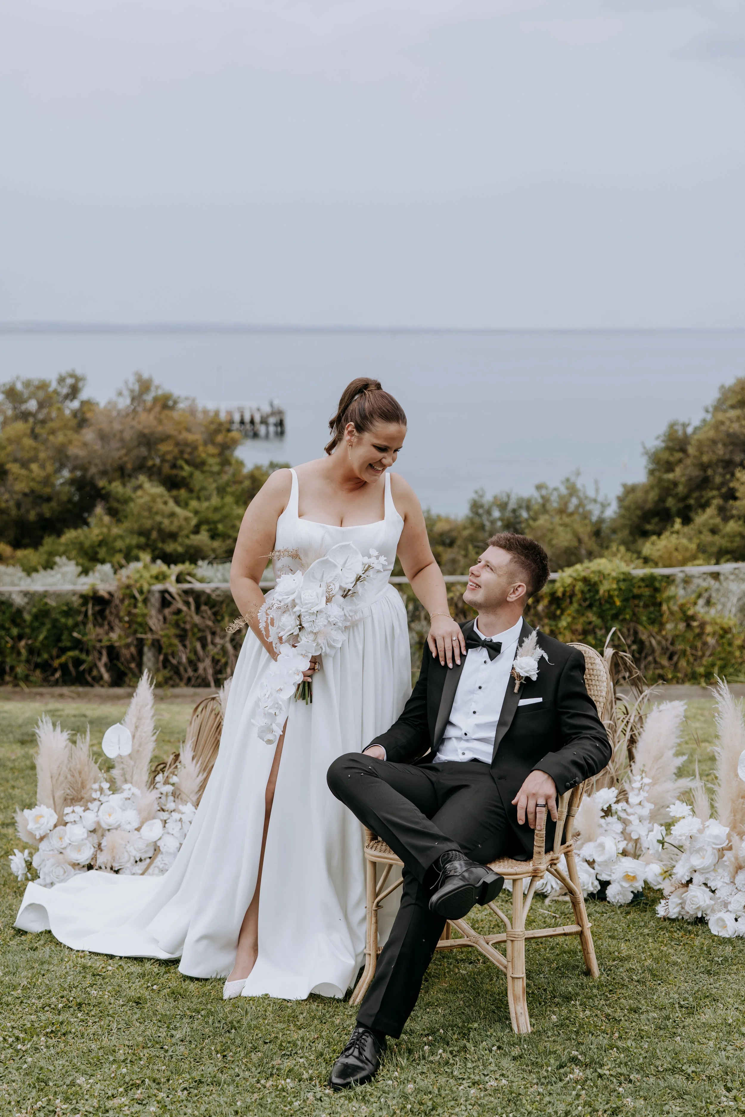 A bride in a white wedding dress and a groom in a black tuxedo with a bow tie are outdoors near a body of water. The bride is holding a large bouquet and smiling at the groom, who is seated on a wicker chair. Floral decorations and pampas grass arrangements are in the foreground, and trees and a pier are in the background.