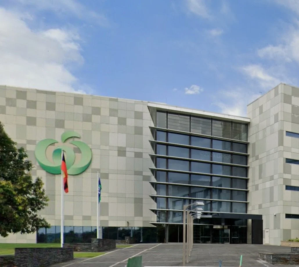 Modern office building with large glass entrance and green logo on facade, surrounded by flags and trees.