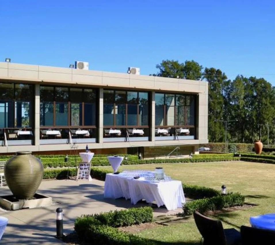 Outdoor event space with tables set with white tablecloths, modern building in the background, and a large decorative vase on a patio.