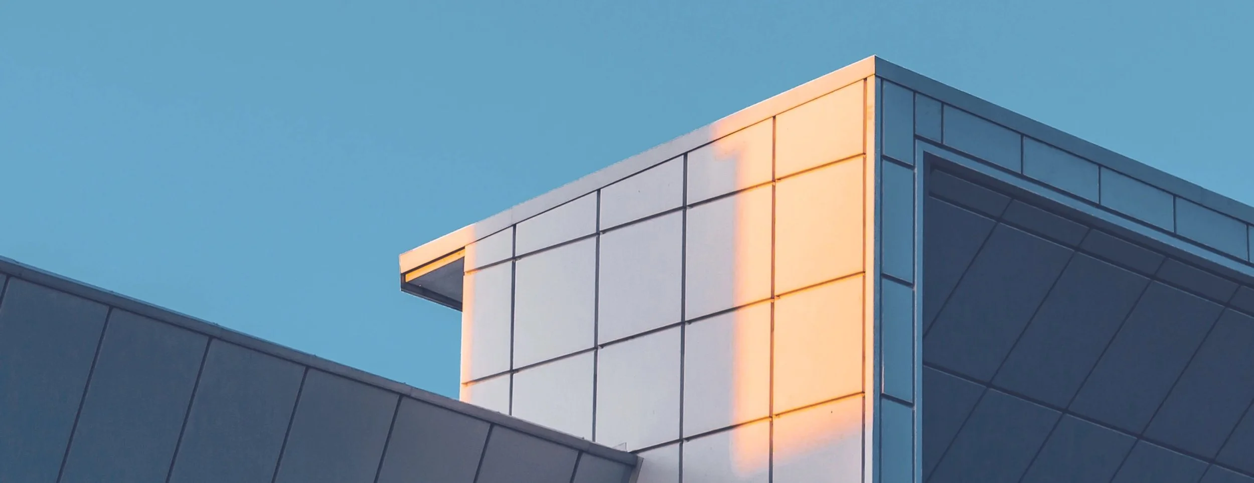Modern building with geometric design, reflecting sunlight, against a clear blue sky.