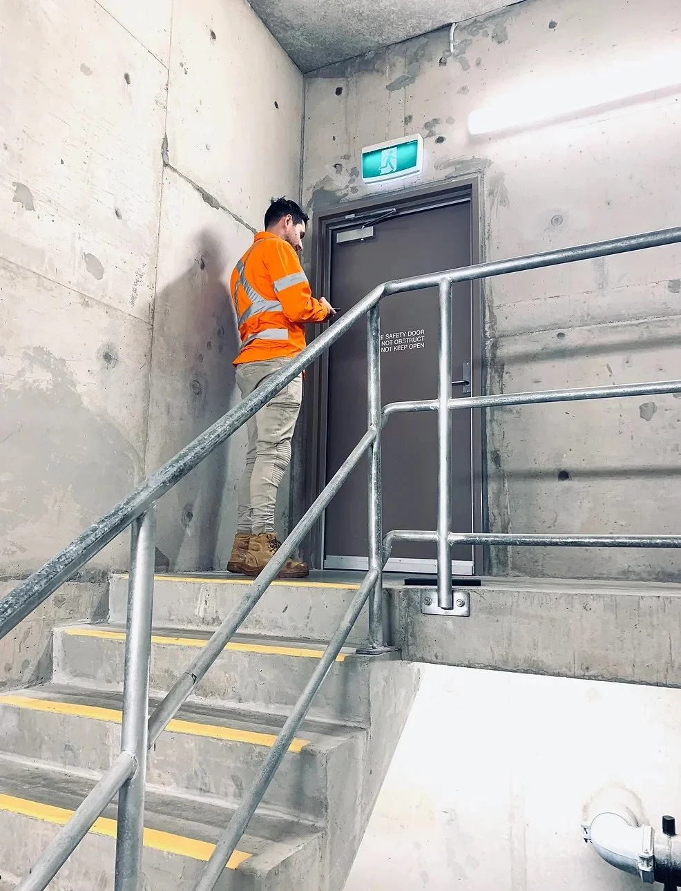 A person in an orange safety vest standing on stairs next to a safety door in a concrete building.