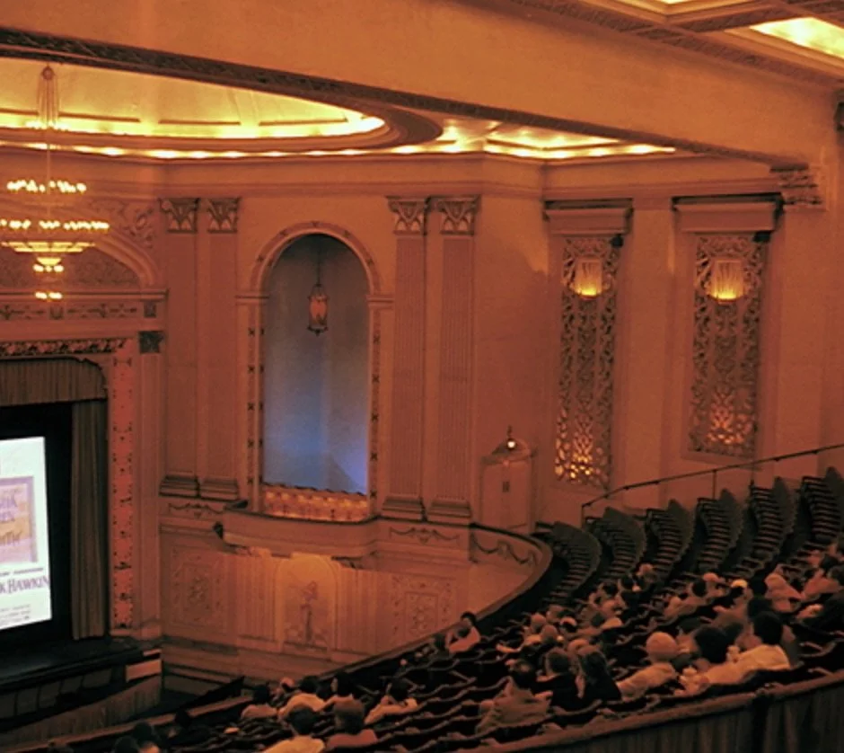 Interior of a large, ornate auditorium with people seated and a screen on stage.