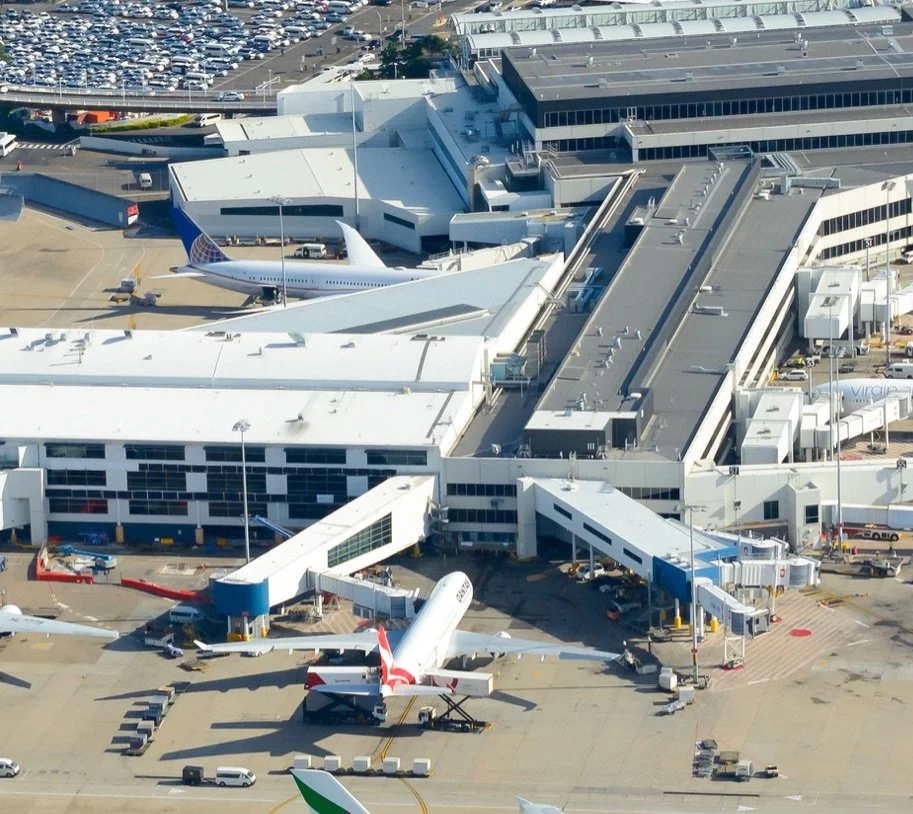 Aerial view of an airport terminal with multiple airplanes docked at gates, surrounded by various airport vehicles and nearby parking lots.