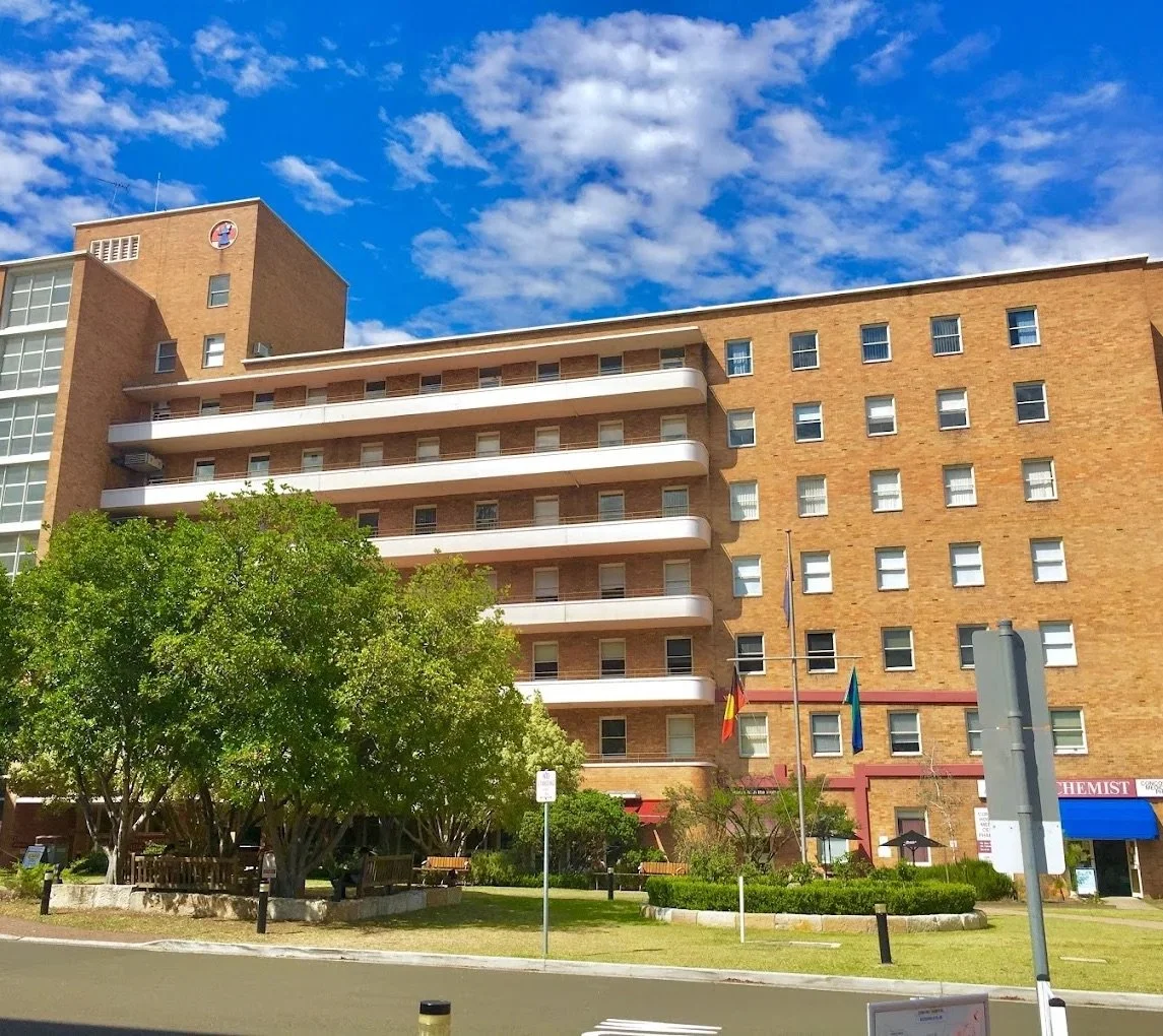 Large brick building with balconies and multiple windows, surrounded by trees and grass under a blue sky with scattered clouds.