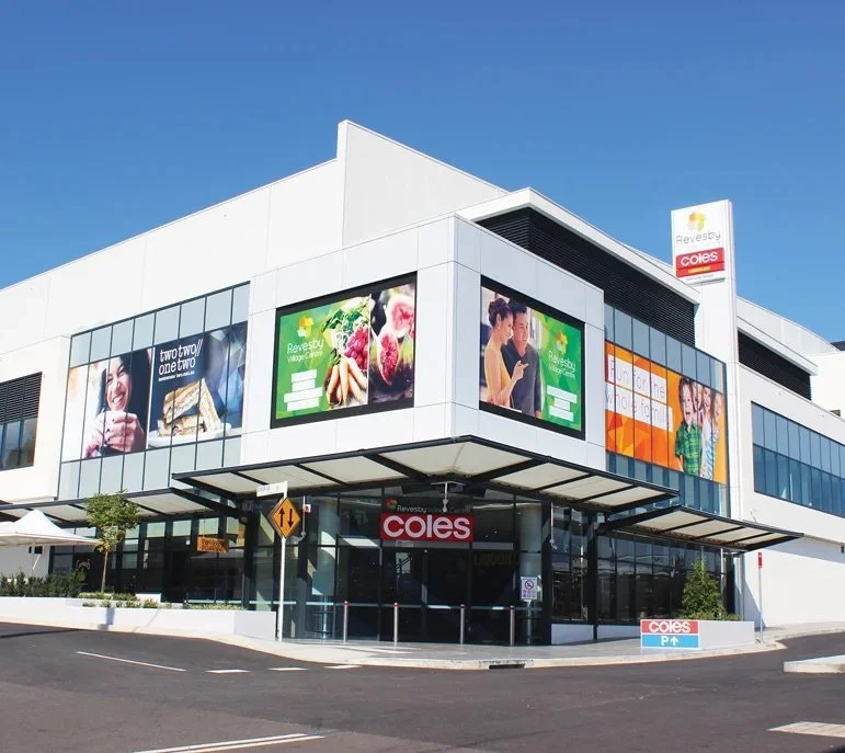 Modern shopping center building with large glass windows and colorful advertisements, featuring a Coles supermarket entrance under a blue sky.