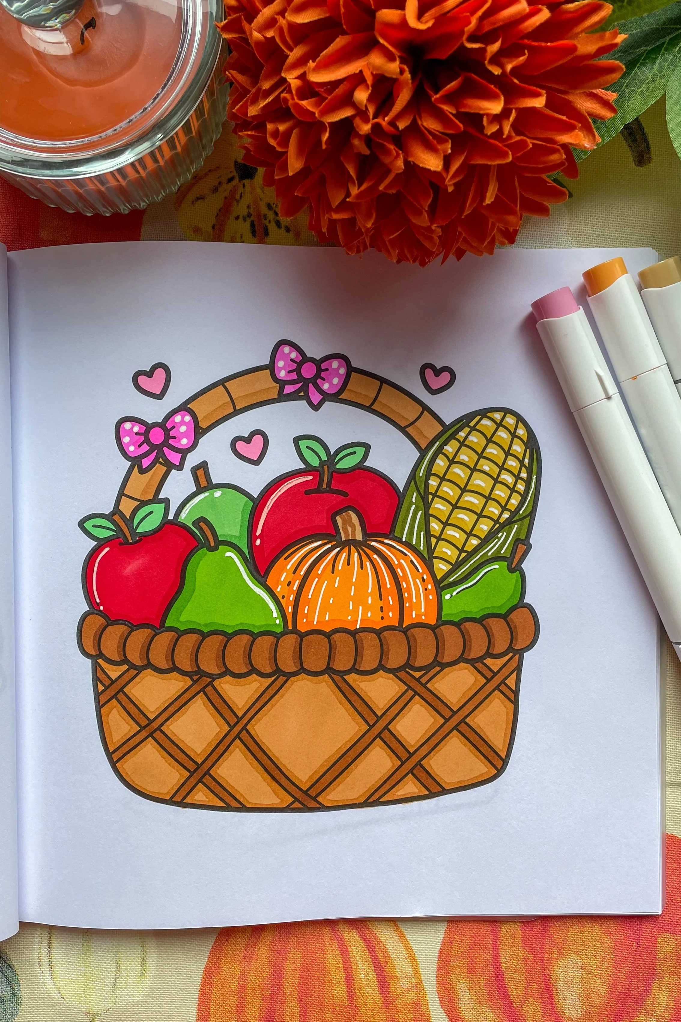 Colorful coloring page of a woven basket filled with pumpkins, apples, corn, and small hearts, decorated with pink bows.