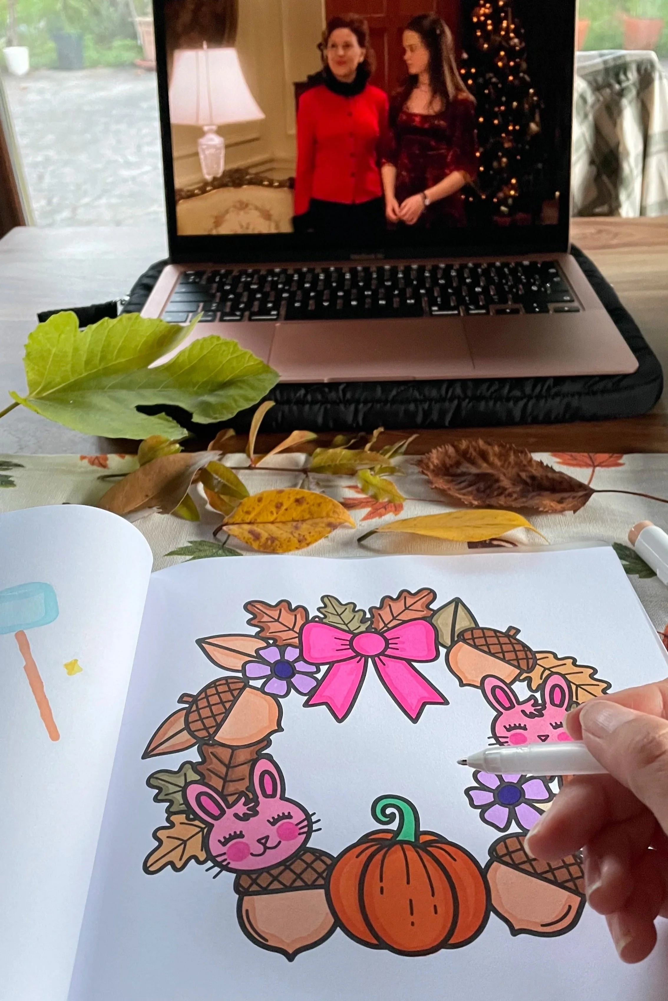 Coloring page with a Halloween theme depicting a pumpkin, a pink bunny, ribbons, fall leaves, flowers, acorns, and a sprig of holly, with a hand coloring it with a marker.